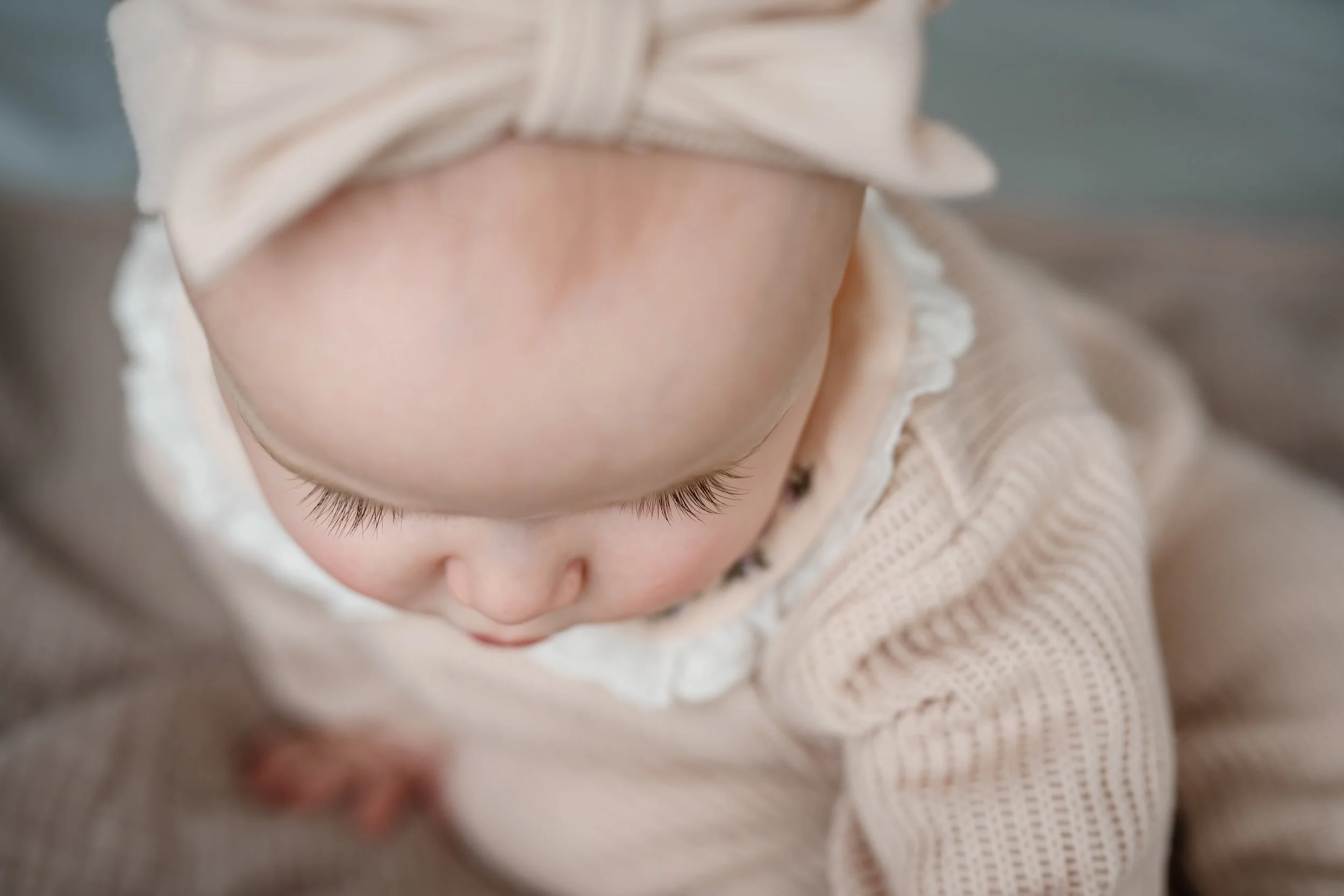 Close-up of a baby wearing a soft knit outfit and bow headband, looking down with delicate eyelashes and gentle natural light.