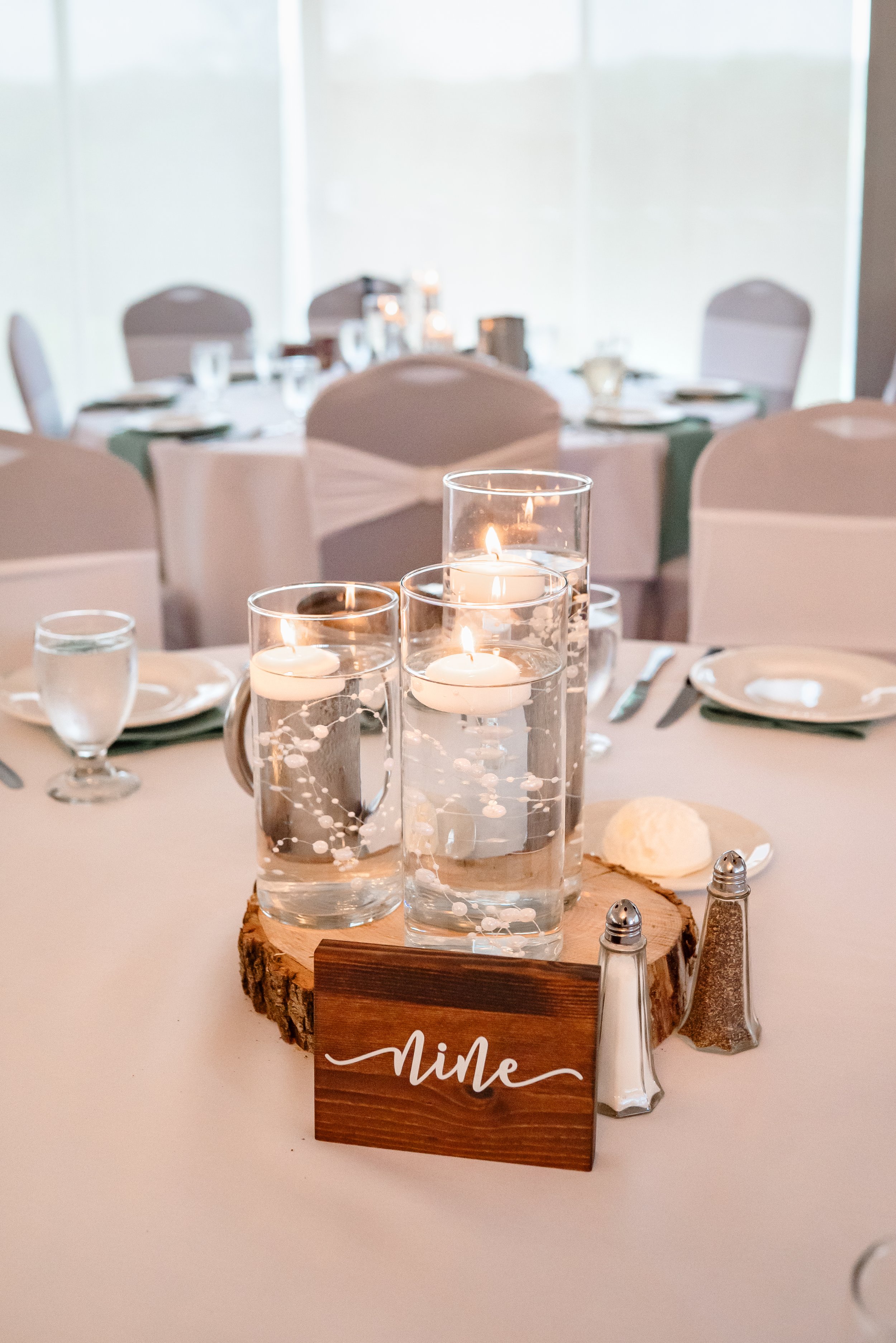 A wedding reception table centerpiece featuring floating candles in glass cylinders, a wooden table number sign, and simple place settings in a softly lit venue.