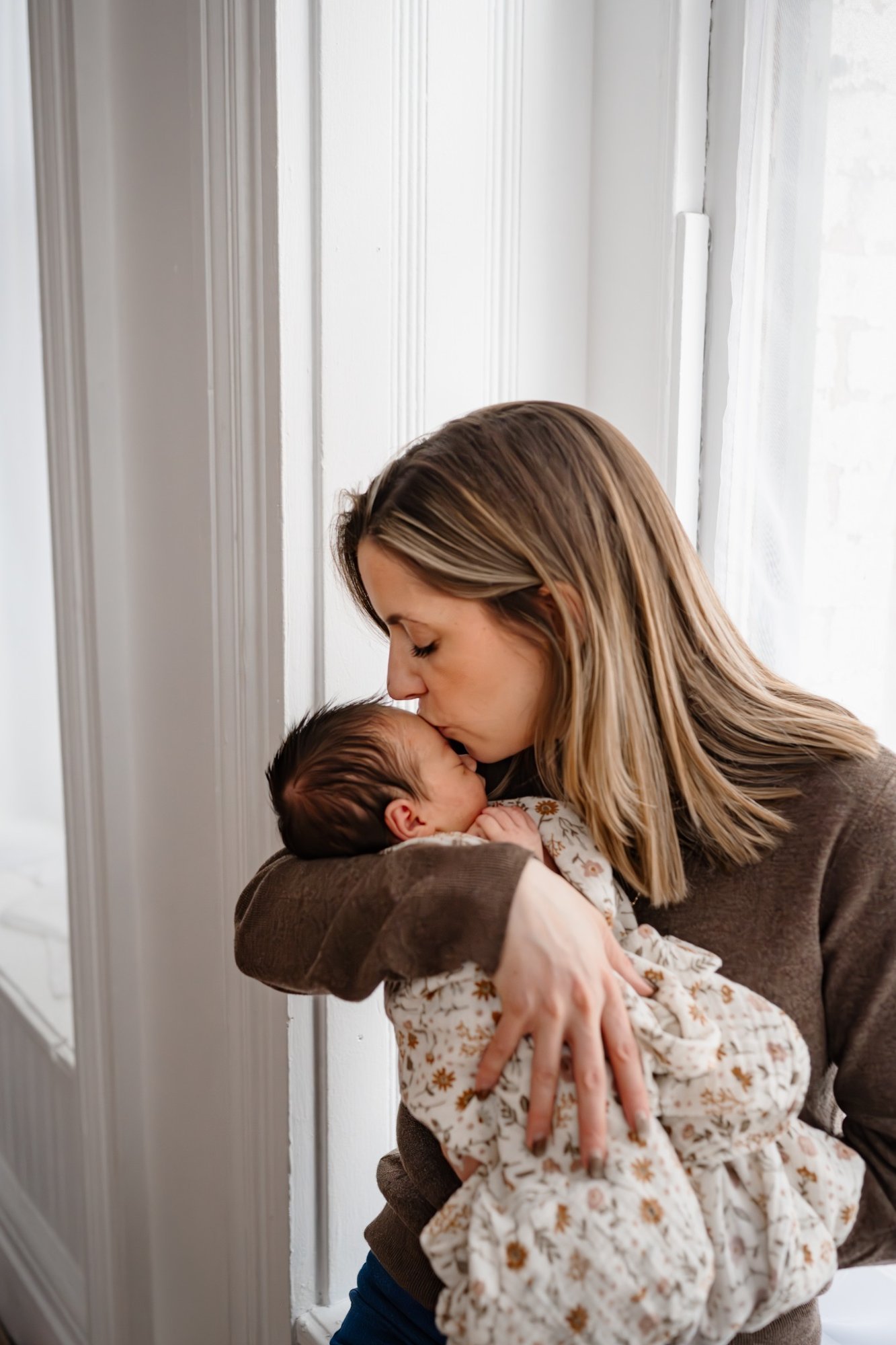 Mother gently kissing her newborn baby while holding them close by a softly lit window during an in-home newborn session.