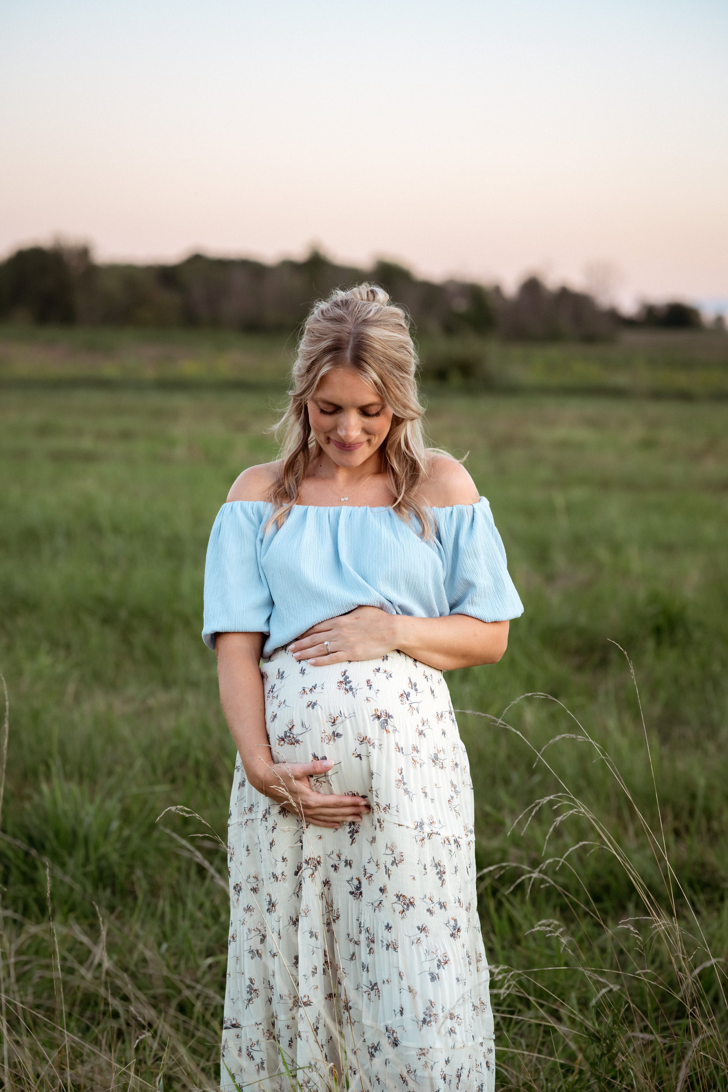 A pregnant woman stands in a grassy field at sunset, gently cradling her baby bump while looking down with a soft smile. Wearing a light blue off-the-shoulder top and a flowing floral skirt, she is surrounded by warm evening light in a peaceful outdo