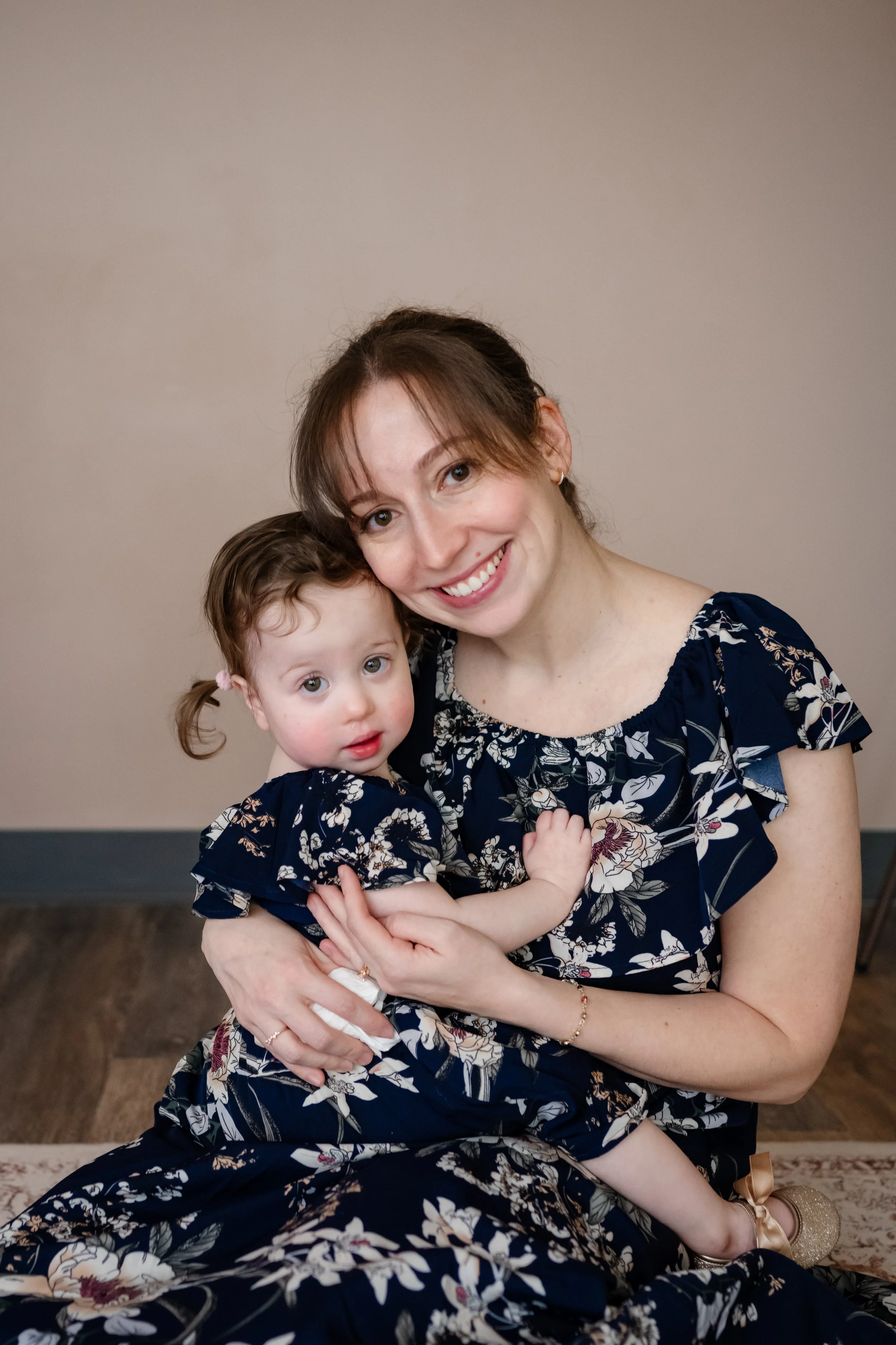 Mother and young daughter sit together in matching navy floral dresses, cuddling close and smiling gently at the camera in a warm indoor setting.