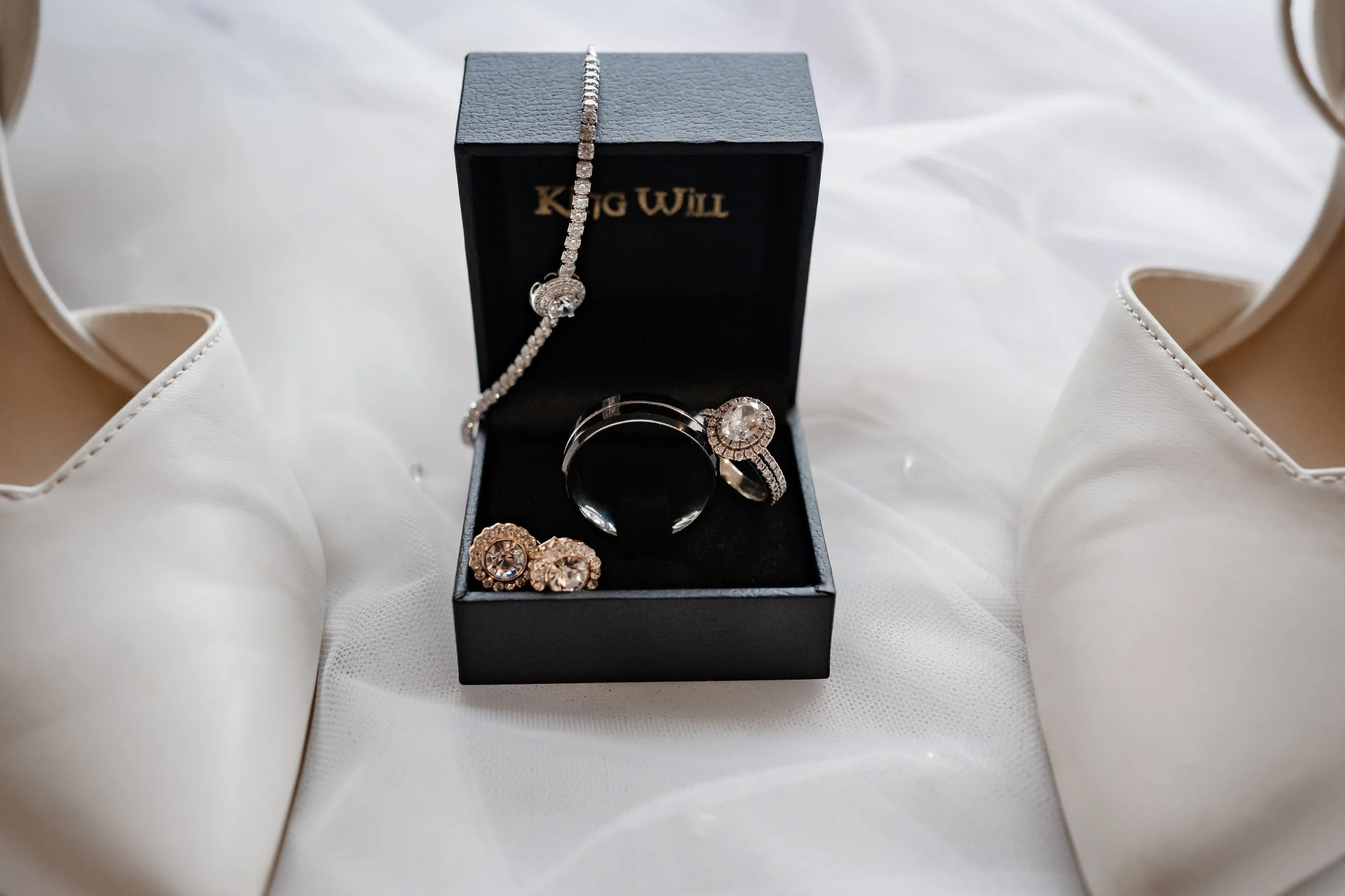 A black ring box displaying wedding rings and jewelry, placed between a pair of white bridal shoes on soft white fabric during wedding detail photography.