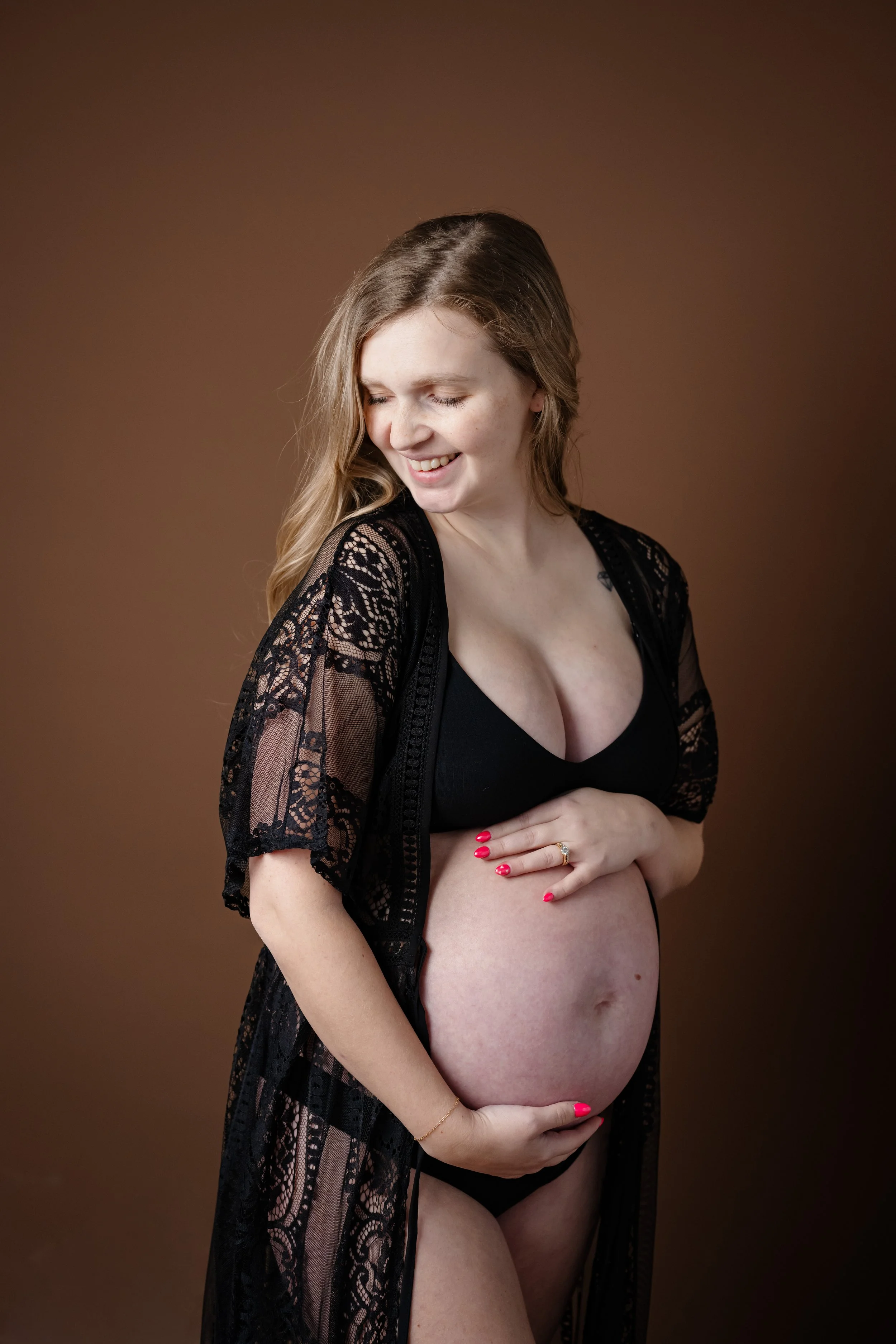 Pregnant woman smiling softly while cradling her baby bump, wearing a black lace robe against a warm brown studio backdrop.
