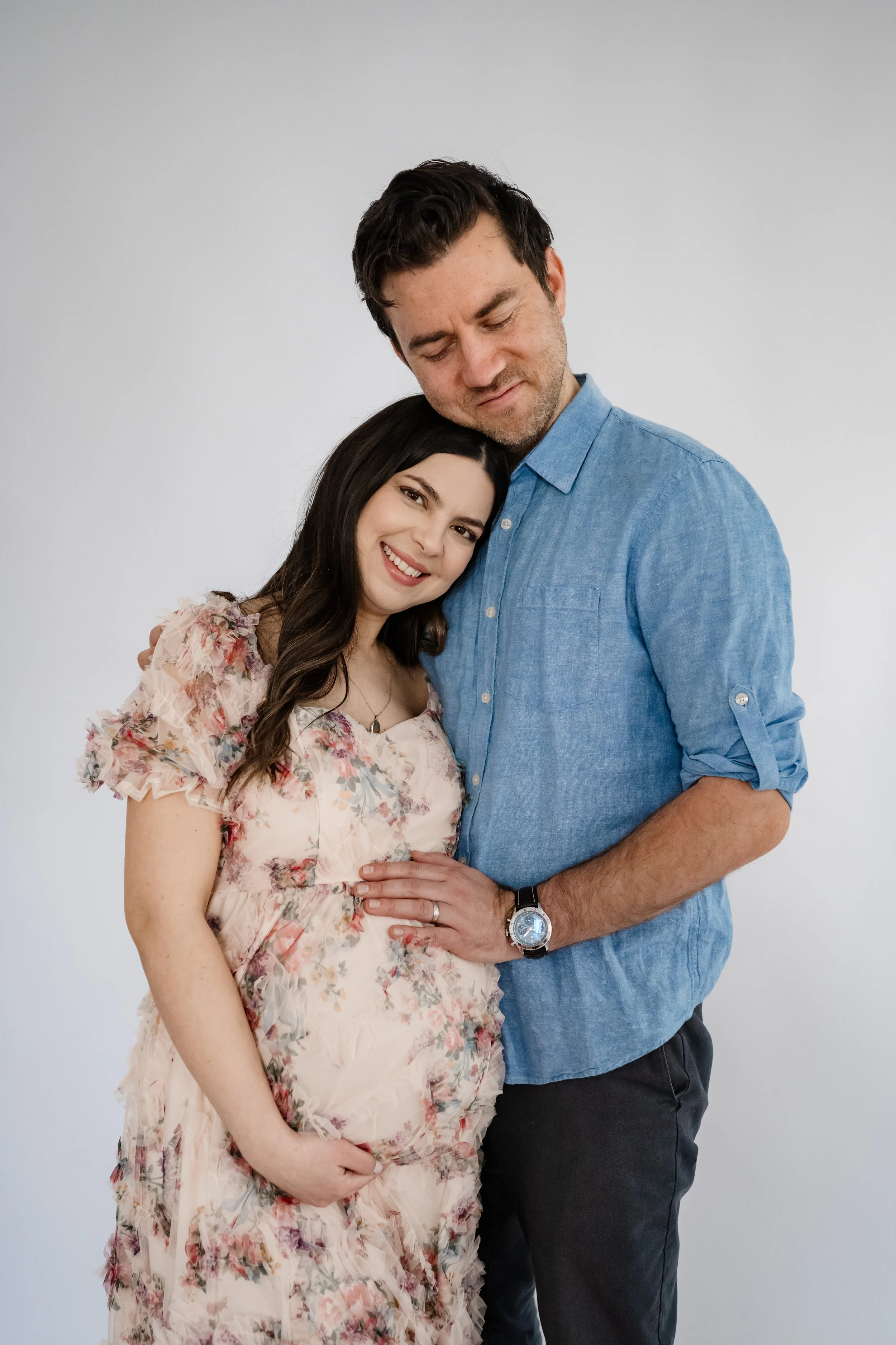 Expectant couple stands closely together against a clean white backdrop, smiling softly as the father rests his hand on the mother’s baby bump, sharing a quiet, intimate moment.