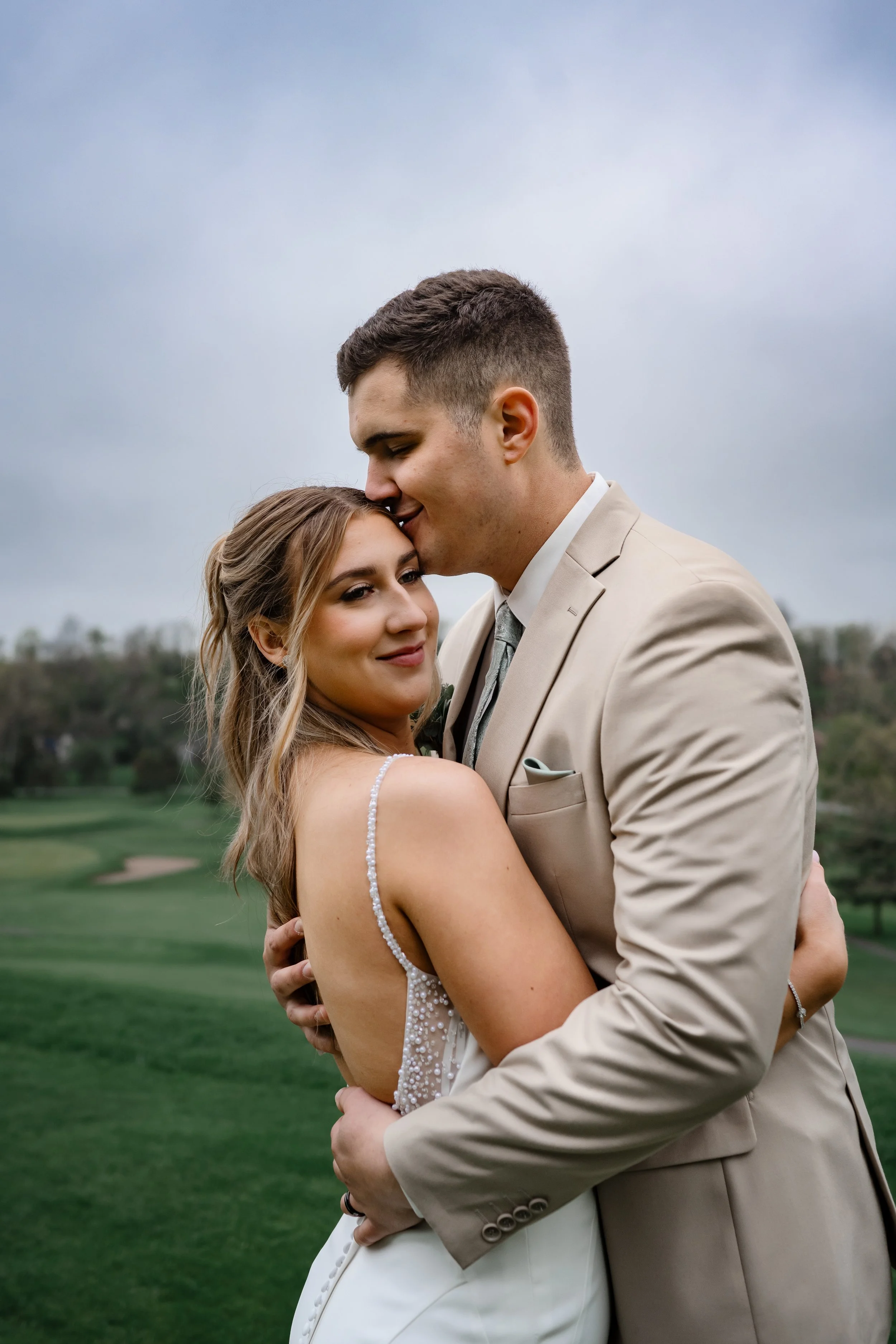 A bride and groom embracing outdoors during a wedding portrait, with the groom kissing the bride’s forehead as they stand on a grassy landscape.