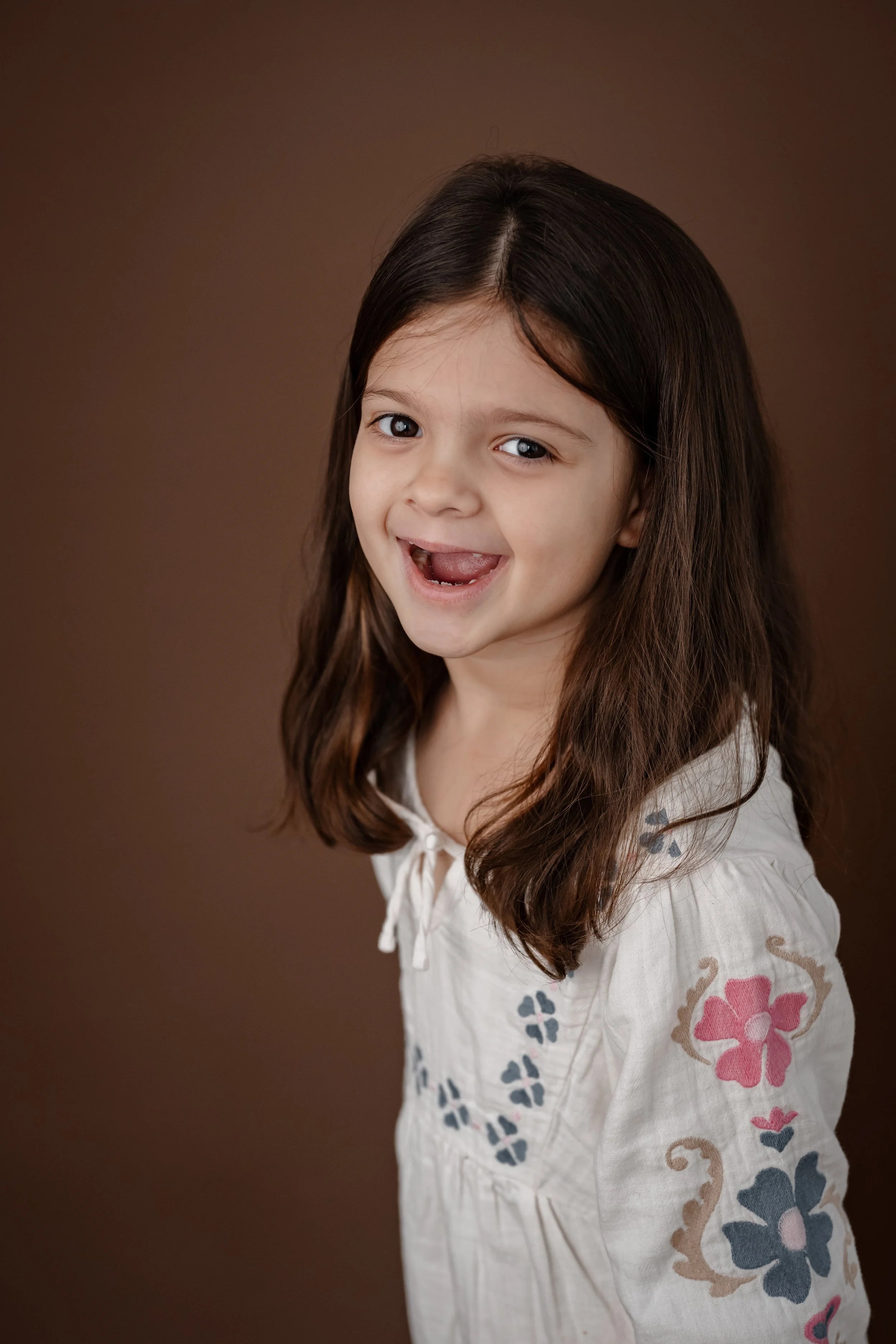 A young girl with long brown hair, smiling and showing a missing front tooth, wearing a white embroidered blouse, against a plain brown background.