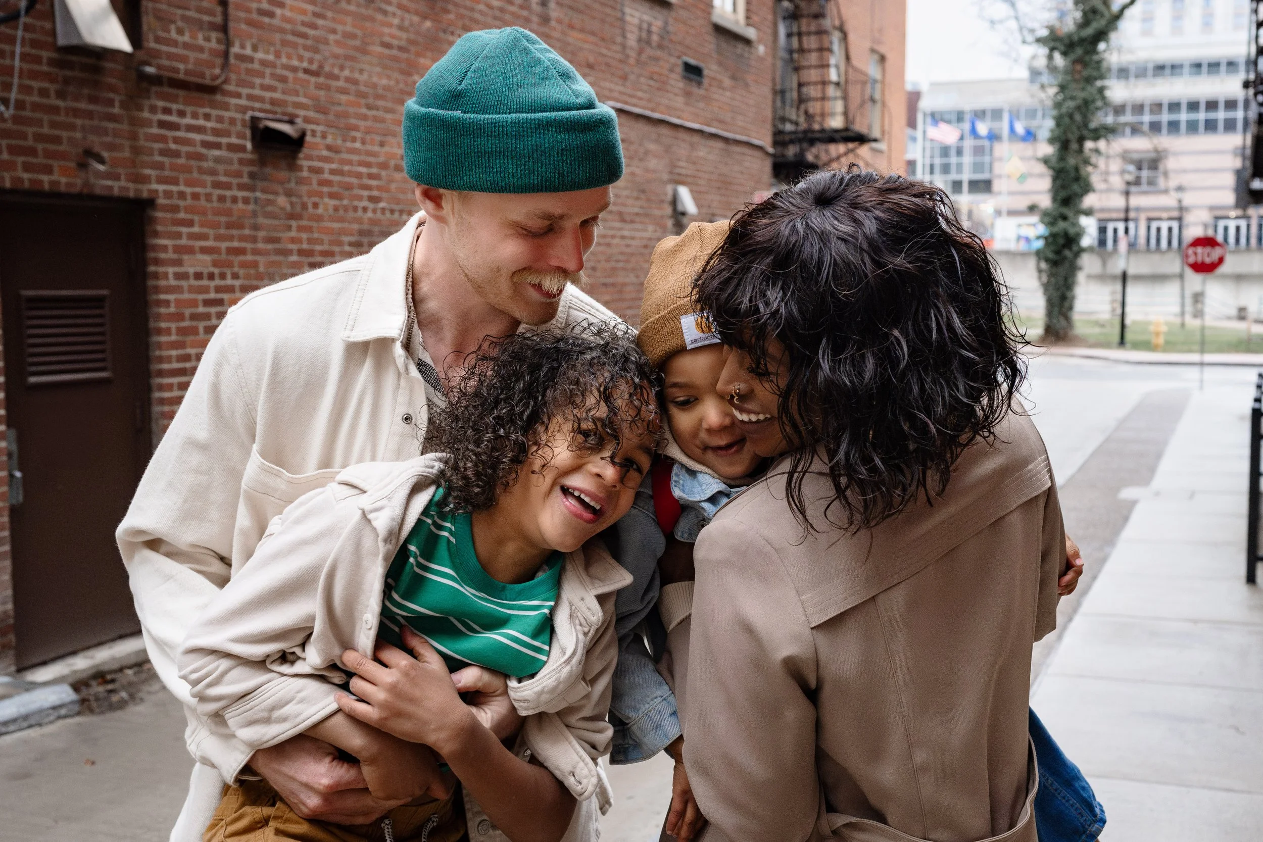A family of four sharing a joyful moment outdoors in an urban setting, with parents holding their two young children close as they smile and laugh together.