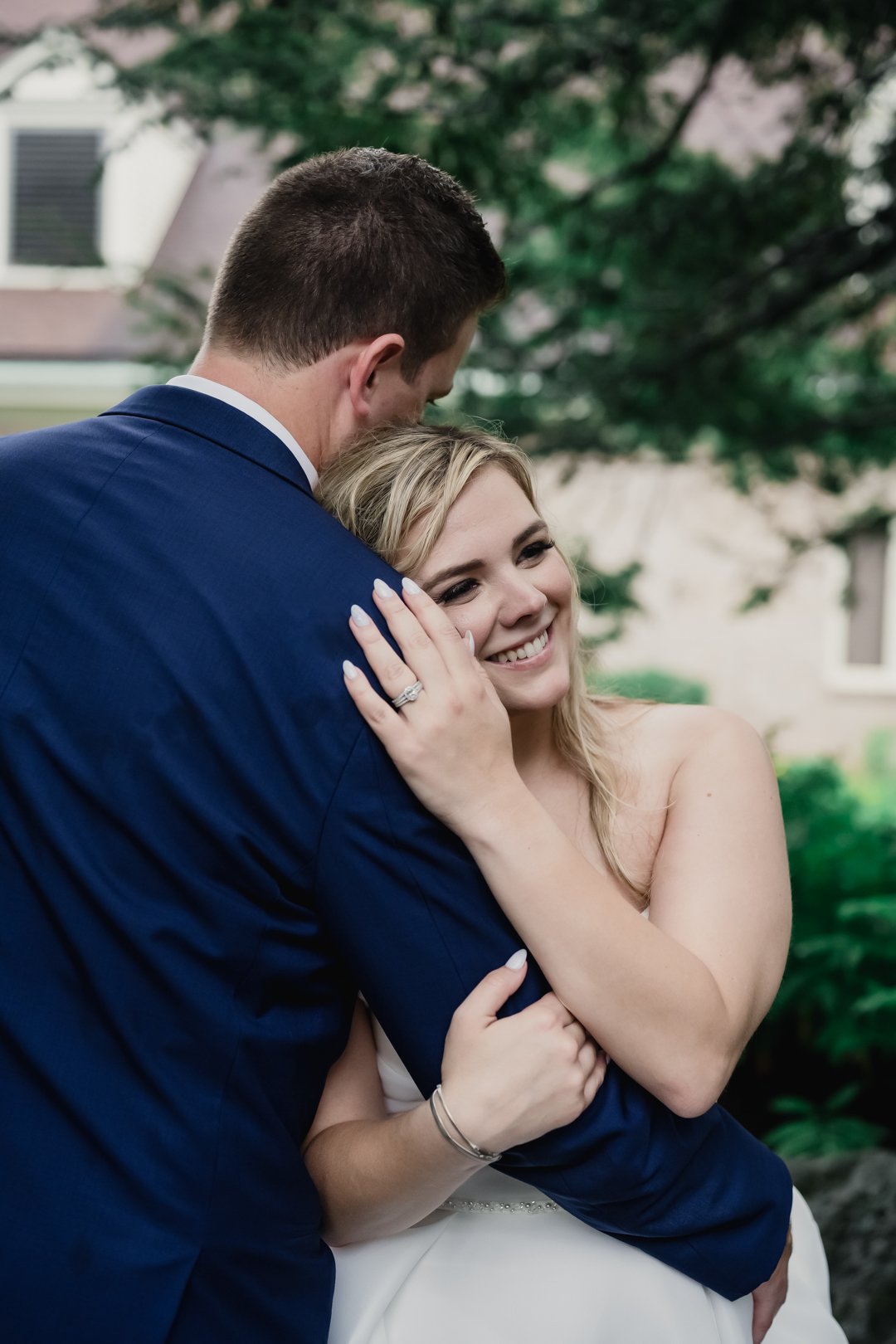 A bride smiling while embracing her groom during an outdoor wedding portrait, resting her head against his shoulder in a quiet, intimate moment.