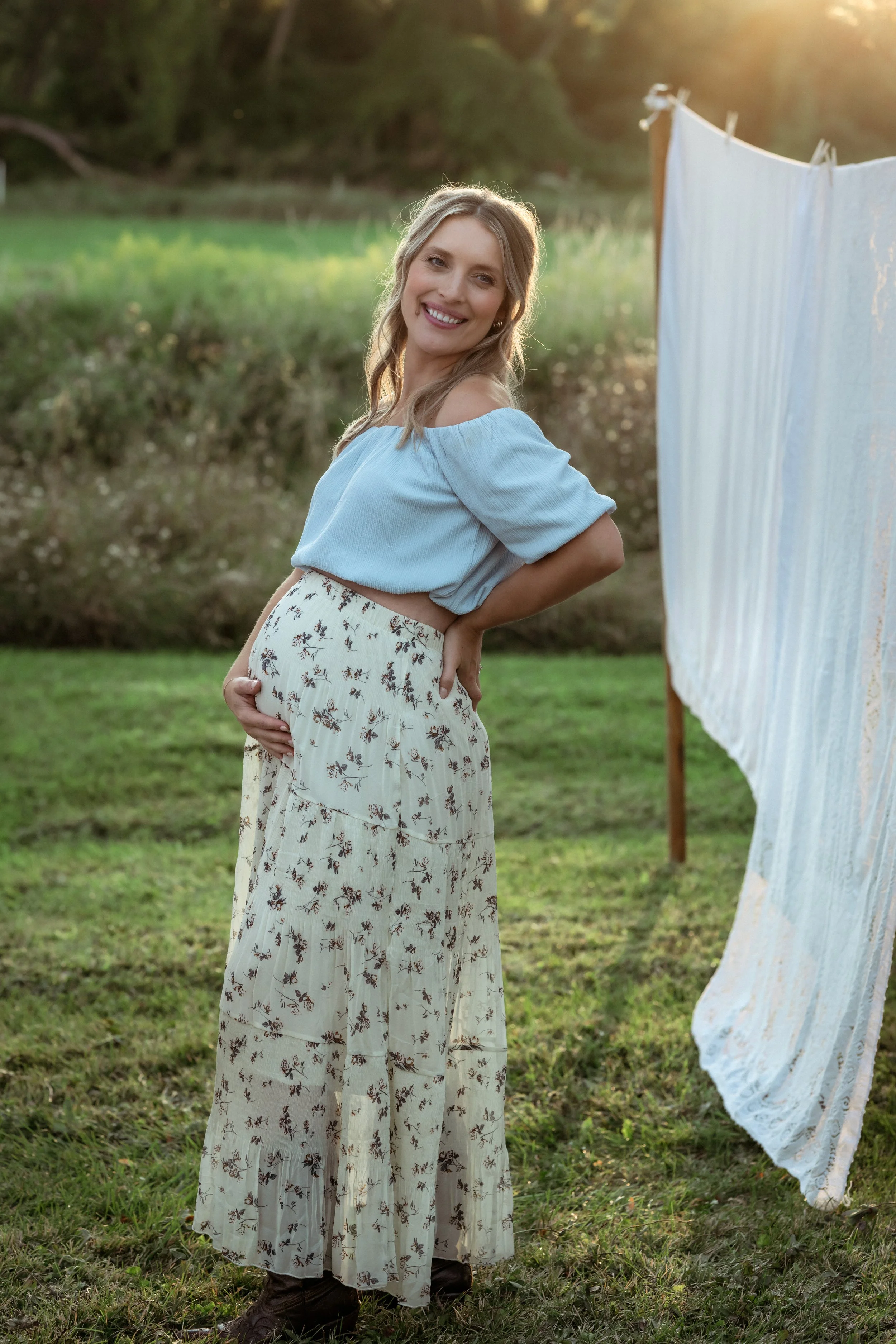 An expectant mother smiling during an outdoor maternity session, standing in a grassy field and gently holding her baby bump, wearing a light blue off-the-shoulder top and a flowing floral skirt in soft evening light.