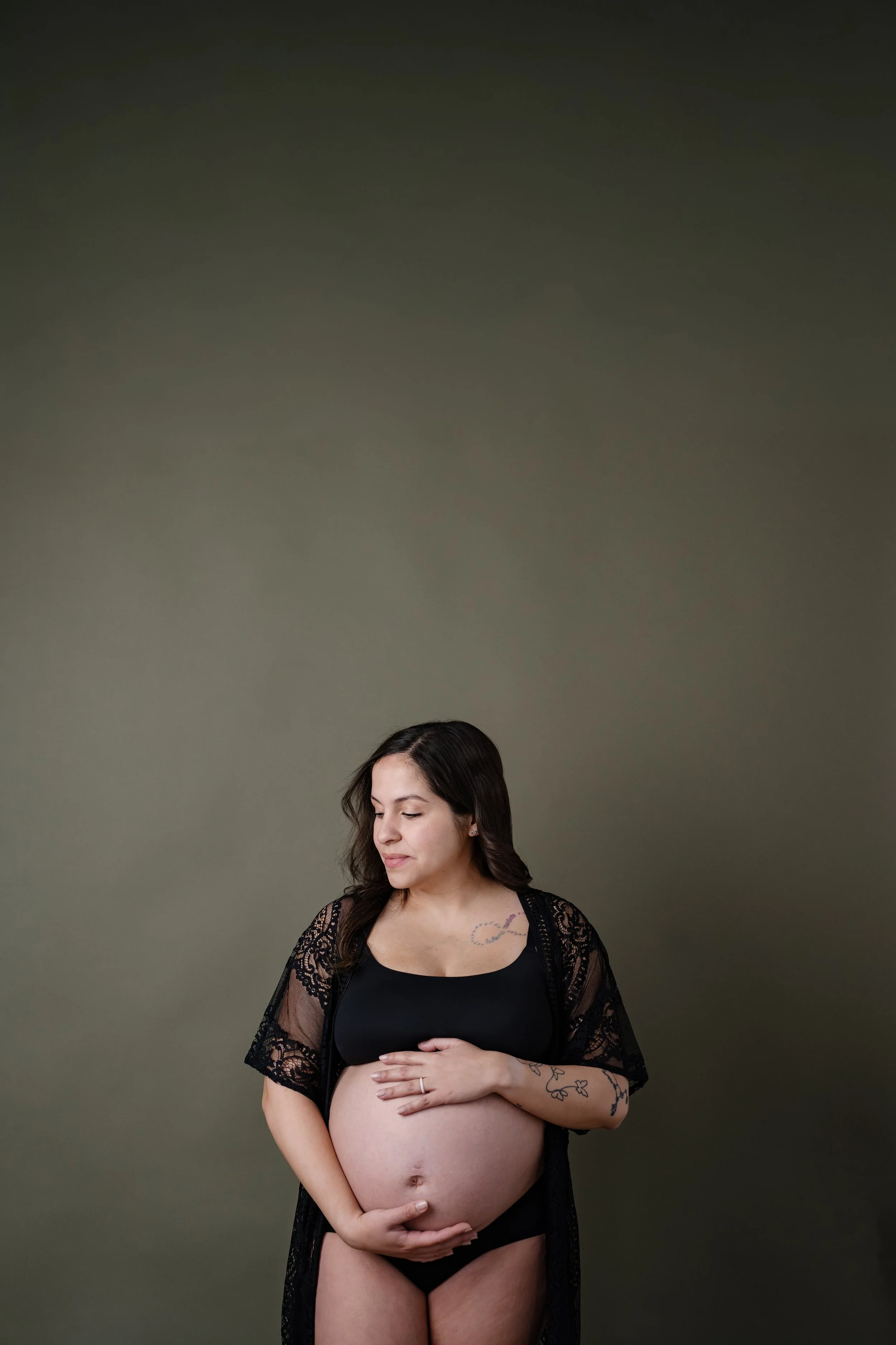 Pregnant woman standing against a muted green backdrop, gently holding her baby bump while wearing a black lace robe and looking down with a calm expression.