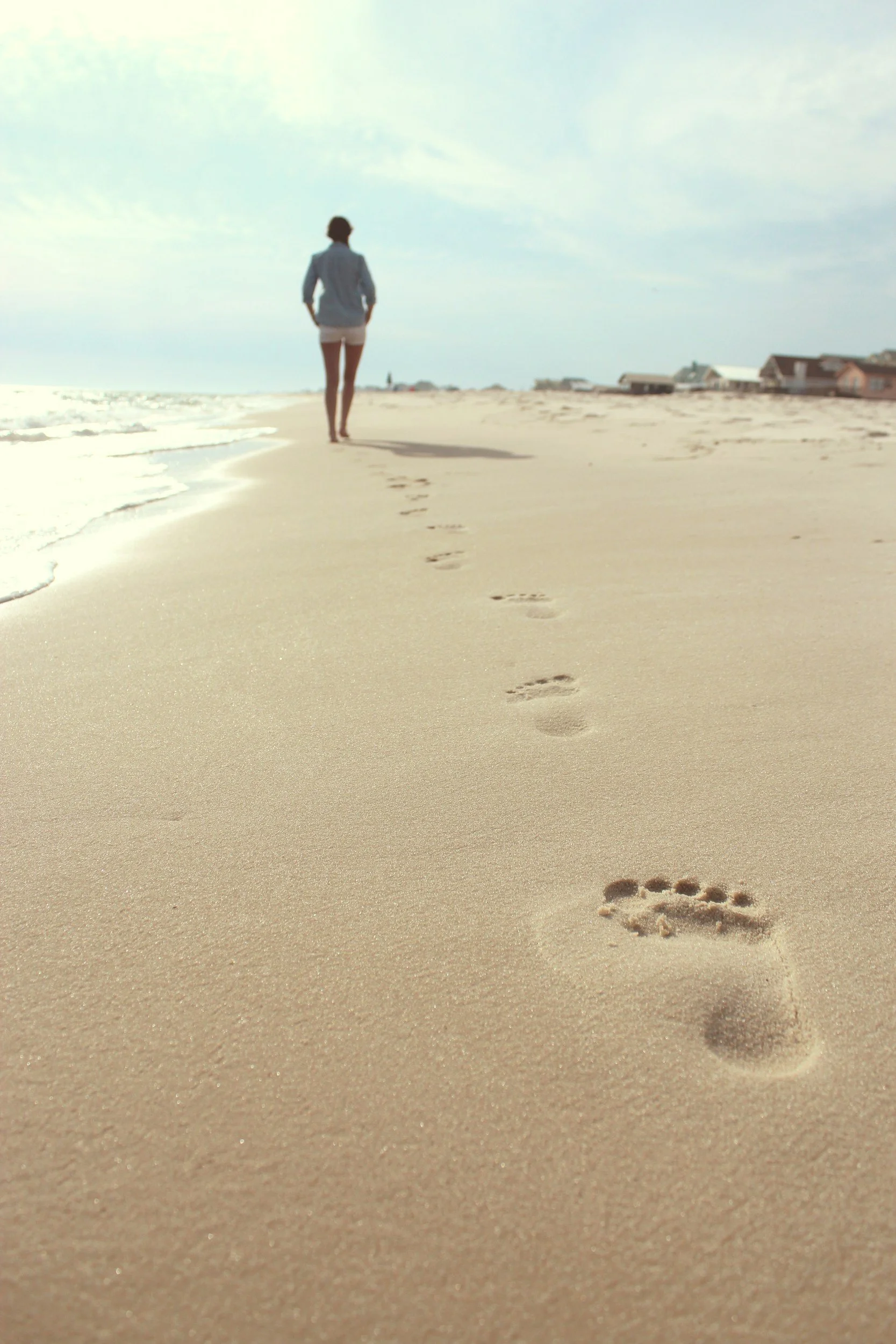 A person walking along a sandy beach with footprints in the foreground and houses in the background under a partly cloudy sky.