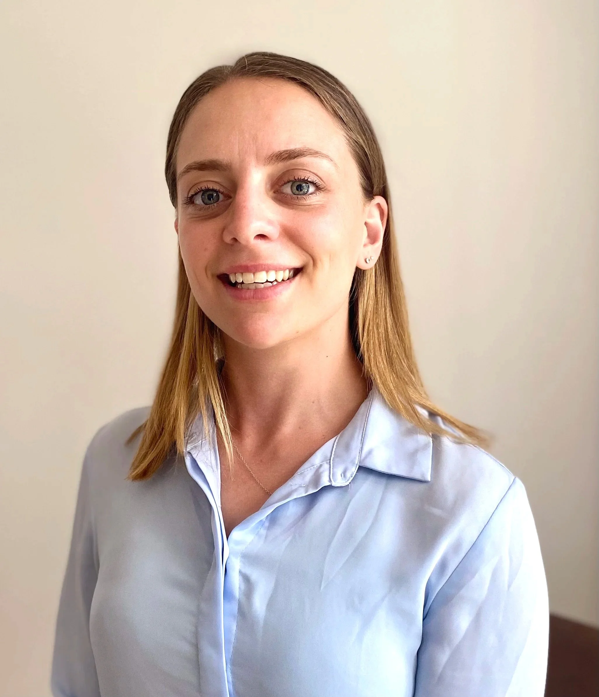 A woman with shoulder-length light brown hair, blue eyes, and fair skin, smiling and wearing a white button-up shirt, standing against a plain light background.