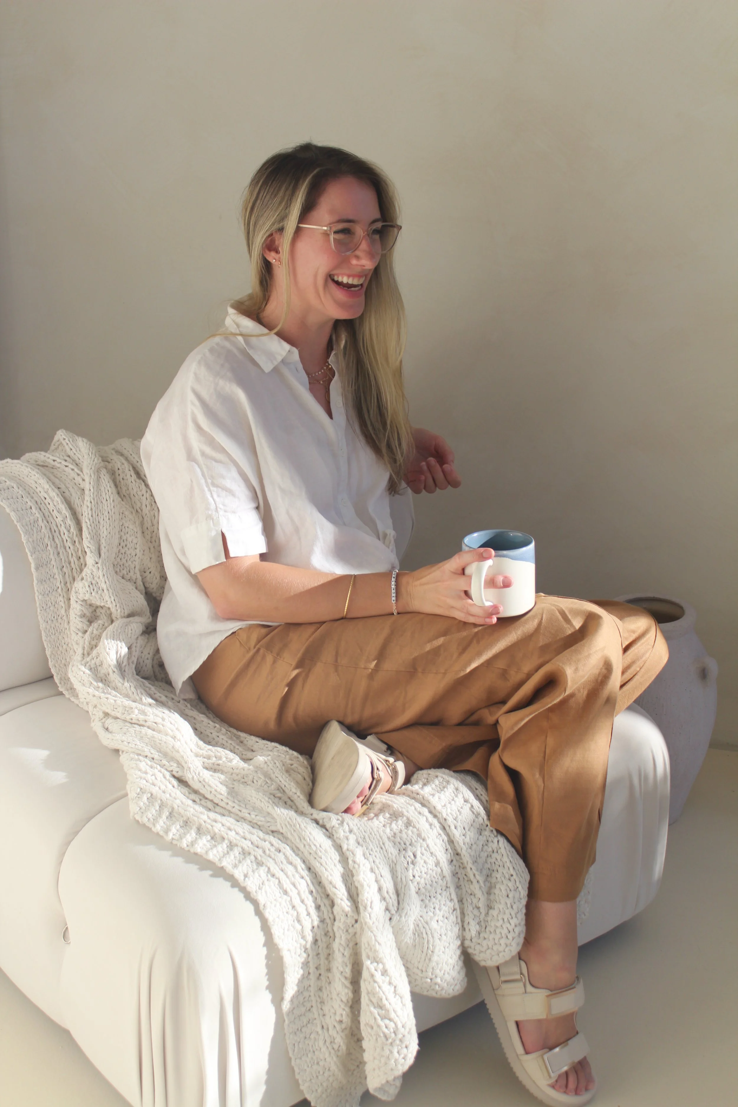 A young woman sitting on a white couch, smiling while holding a ceramic mug, wearing glasses, a white blouse, tan pants, and white shoes, with a cream-colored knit blanket on the couch.