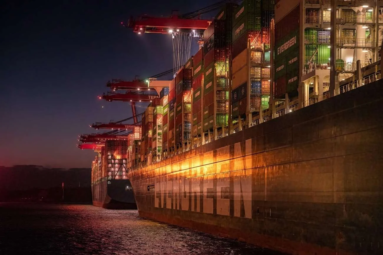Large cargo ships docked at a port during nighttime, with cranes extending over the ships, illuminated by orange and white lights.