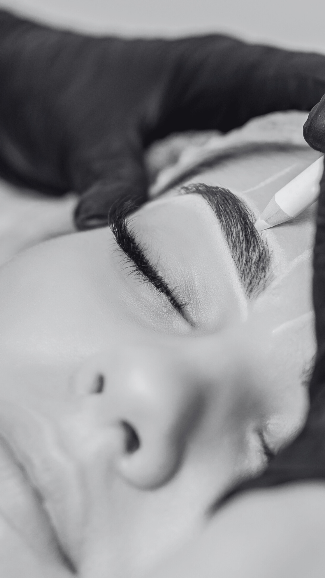 Close-up of a person with their eyes closed getting their eyebrows shaped or tattooed, with the artist's hand holding a pencil or tool, in black and white.