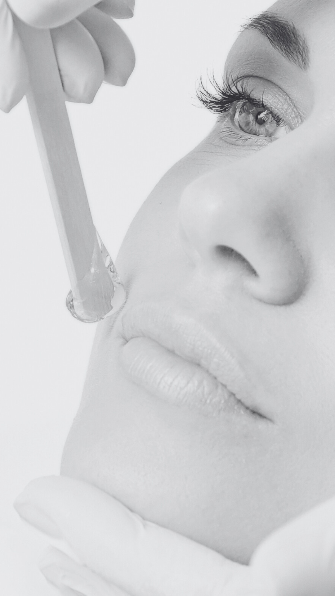 Close-up black-and-white photo of a woman's face with a dropper, about to apply or drop serum on her face, focusing on her eye, nose, and lips.