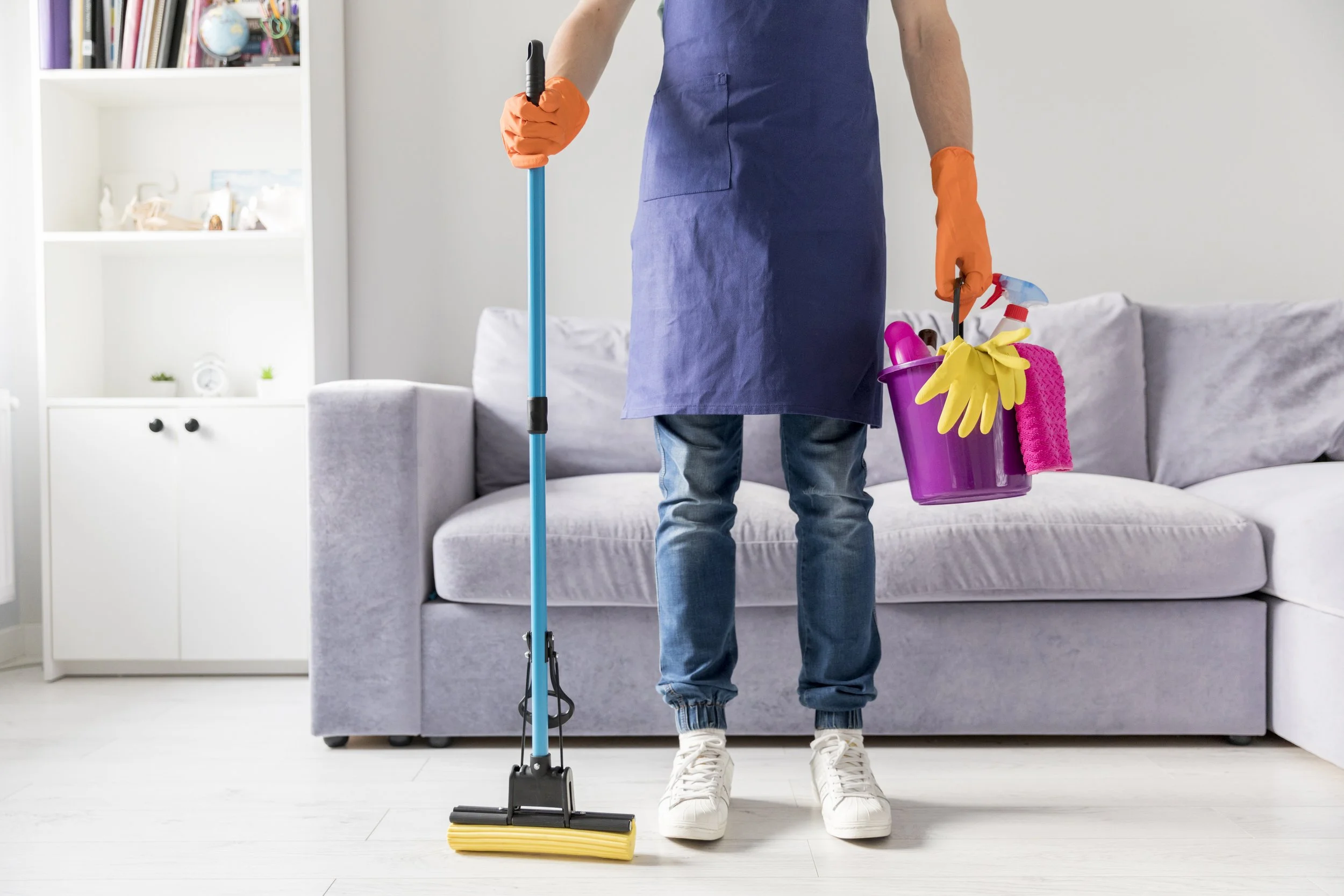 Cleaning staff wearing blue apron and gloves holding cleaning supplies, standing in front of a gray sofa