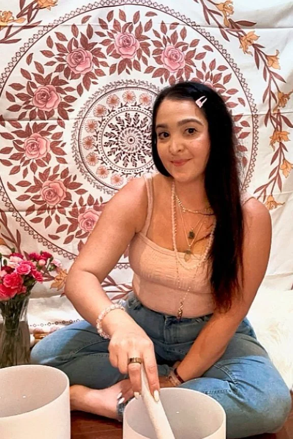 A young woman sitting cross-legged on the floor, holding a white stick or rod, with a tapestry of pink roses and mandala-like patterns in the background, and a vase of pink flowers nearby.