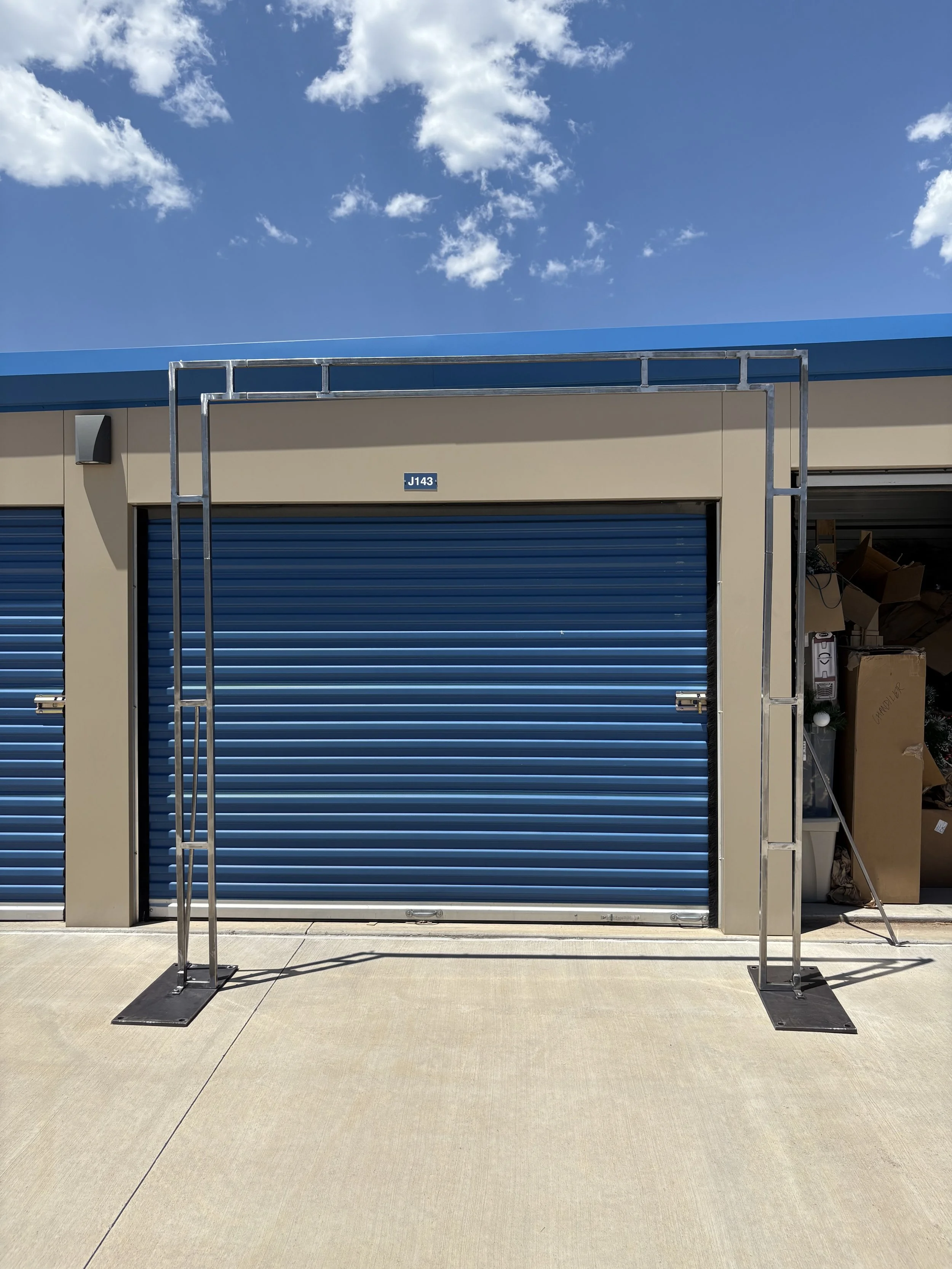 Empty metal frame structure in front of a blue garage door at a storage facility under a partly cloudy sky.