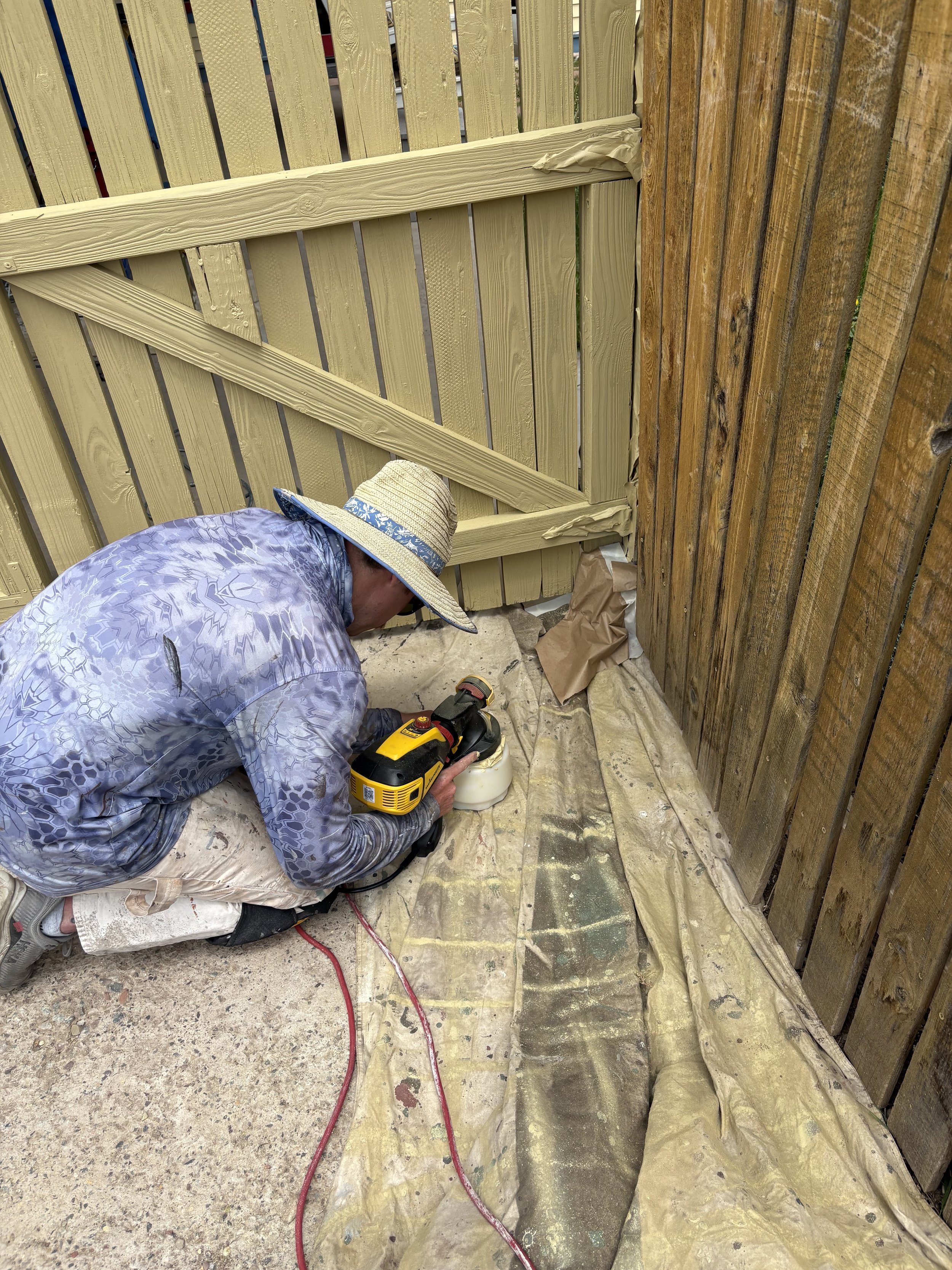 A man kneeling on a tarp applying finishing to a wooden fence with a handheld power sander, wearing a wide-brimmed hat, patterned long-sleeve shirt, and shorts.