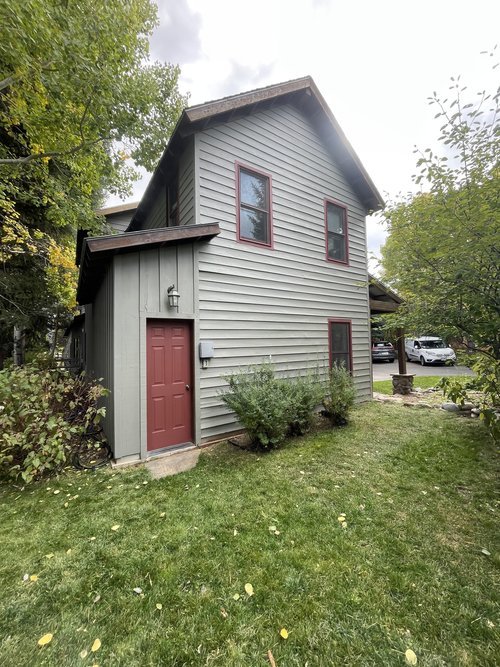 Two-story house with light gray siding, red trim windows, and red front door, surrounded by green grass and trees.