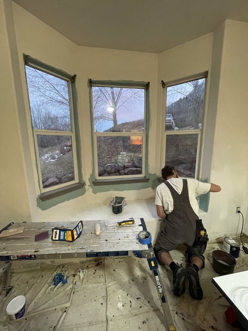 A person installing or repairing three large windows in a room, kneeling on the floor with tools and supplies nearby.
