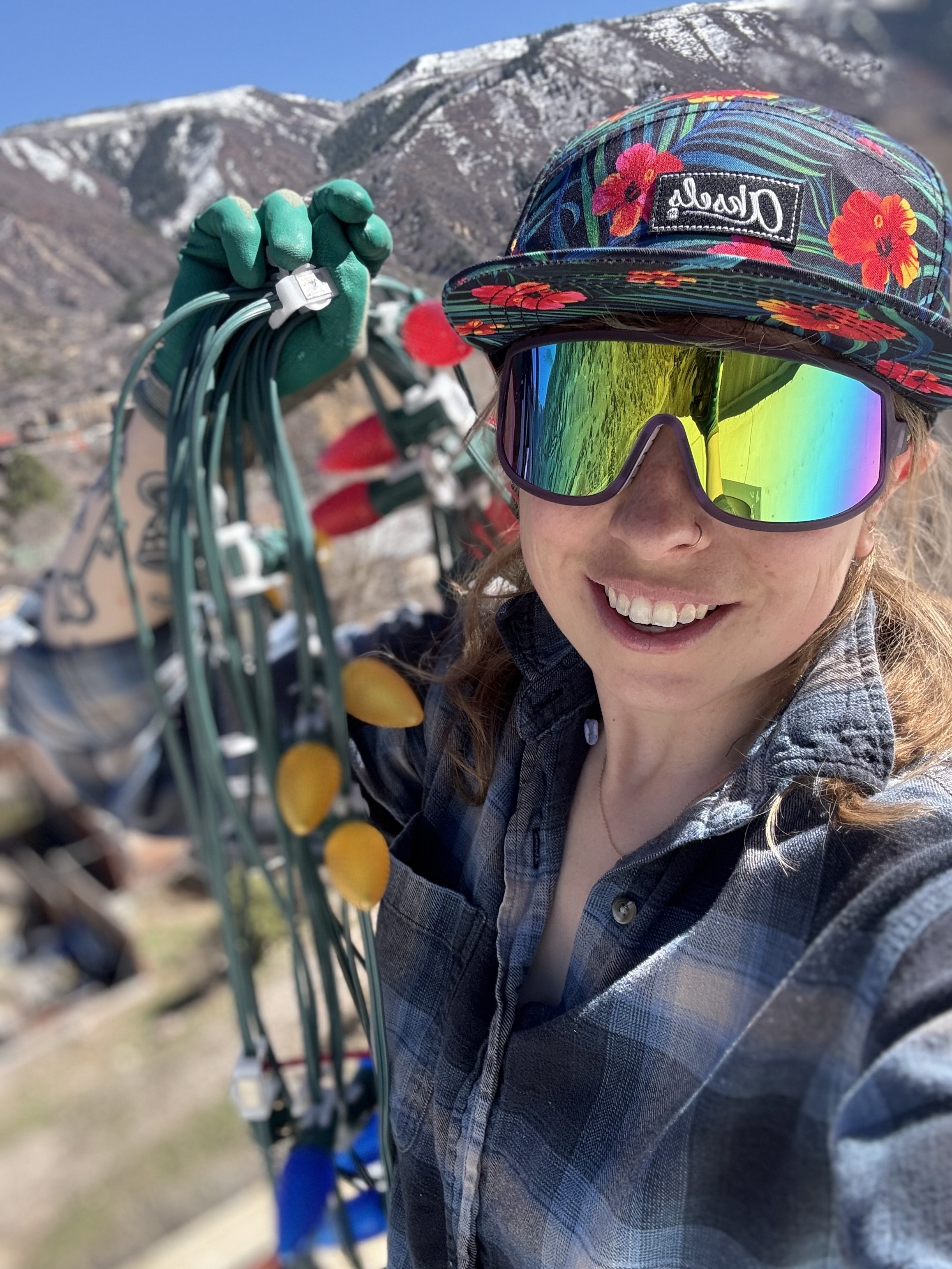 A smiling woman taking a selfie outdoors with a mountain in the background. She is wearing a colorful floral cap, reflective sunglasses, a checkered shirt, and green gloves, and is holding a bundle of electrical cords.