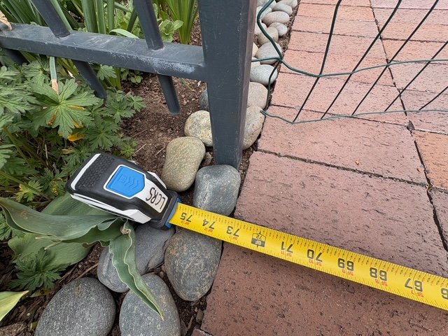 Measuring tape extended along brick pathway near plants and a metal fence.