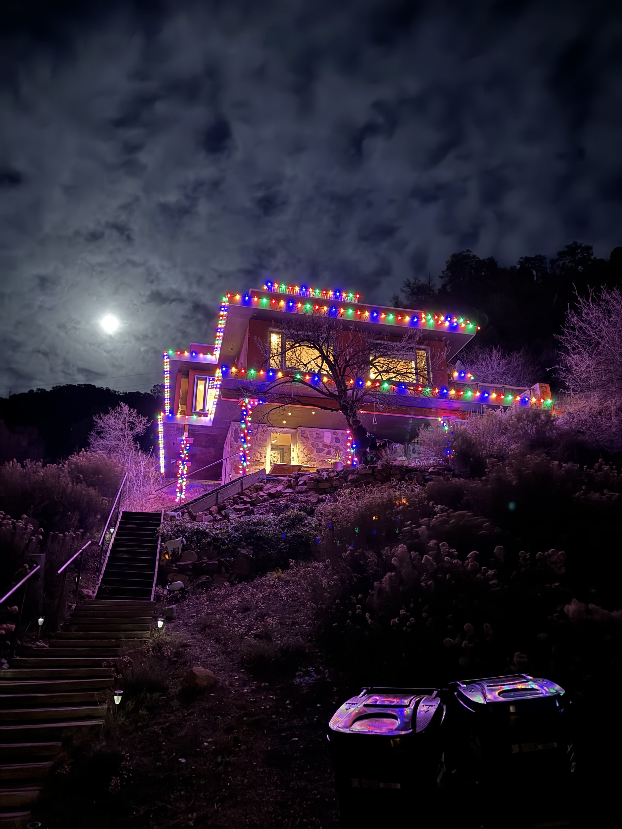 A house decorated with multicolored Christmas lights on the roofline and outer edges, illuminated against a night sky with the full moon and clouds. The house is situated on a hillside, with a staircase leading up to it, and surrounded by trees and p