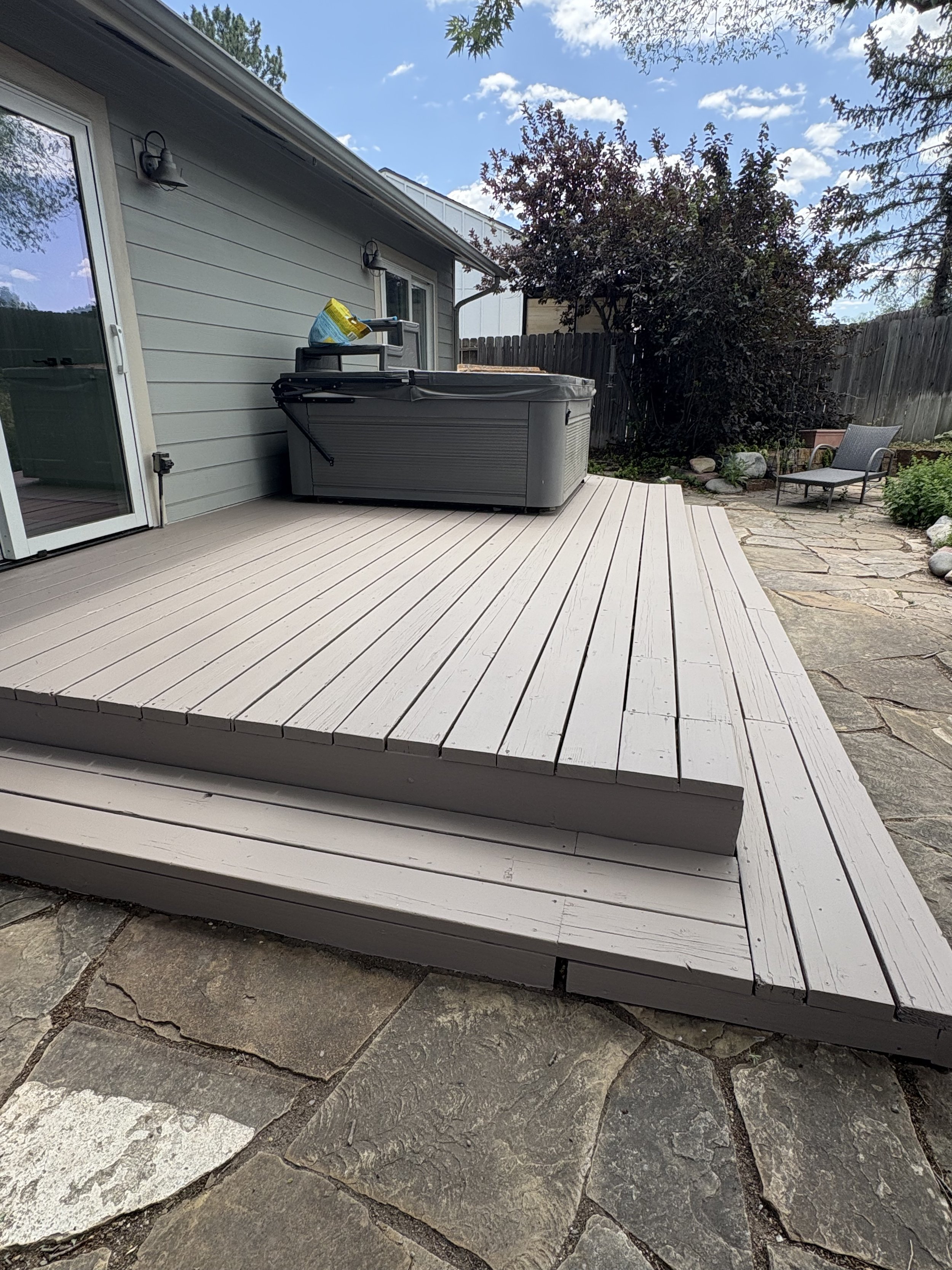 A backyard patio with light gray wooden decking, a hot tub, patio furniture, and a stone pathway with a garden in the background.