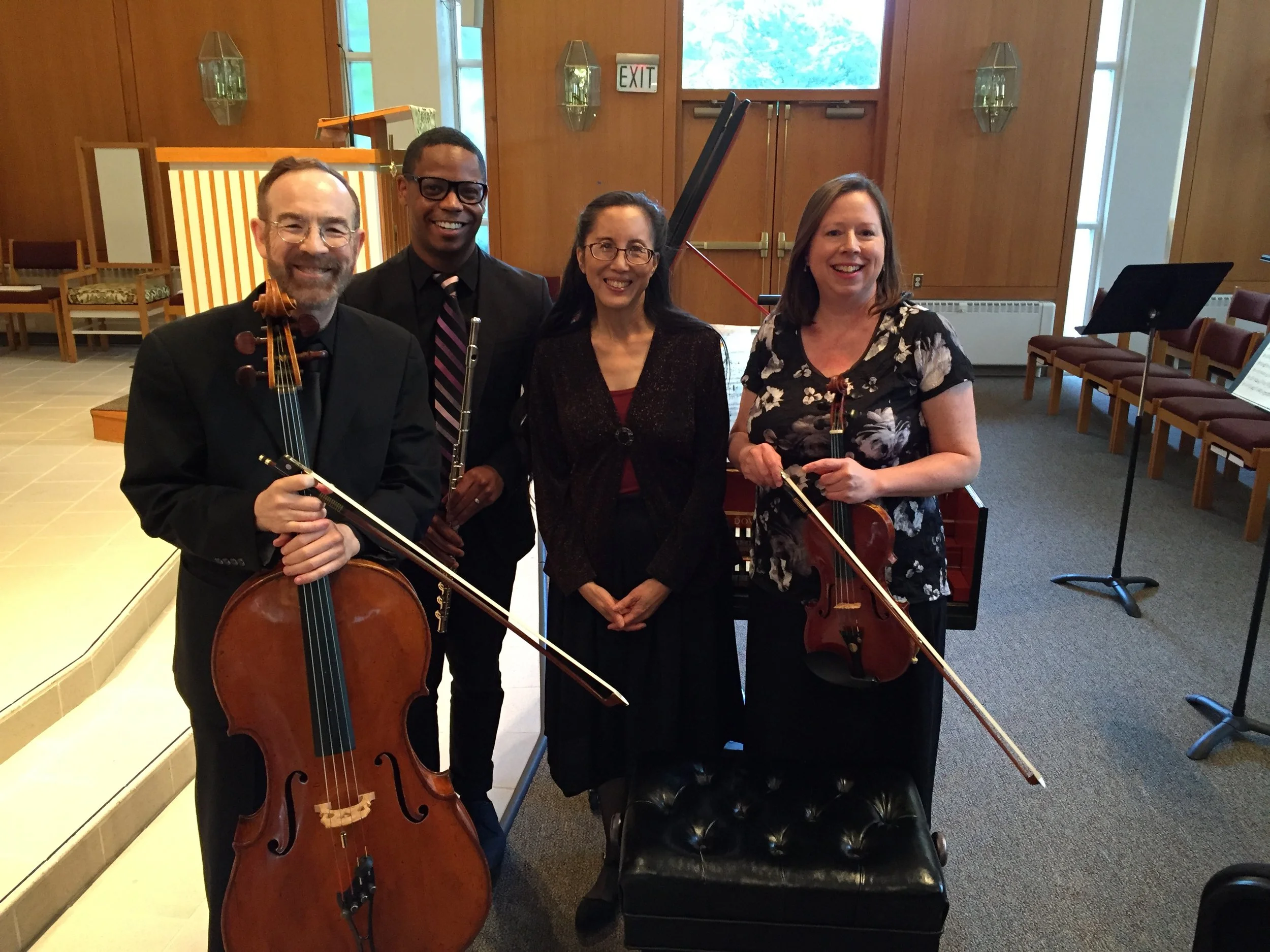 Richmond Chamber Players. Four musicians standing in a room with wooden walls, two holding string instruments and in front of music stands, smiling at the camera.