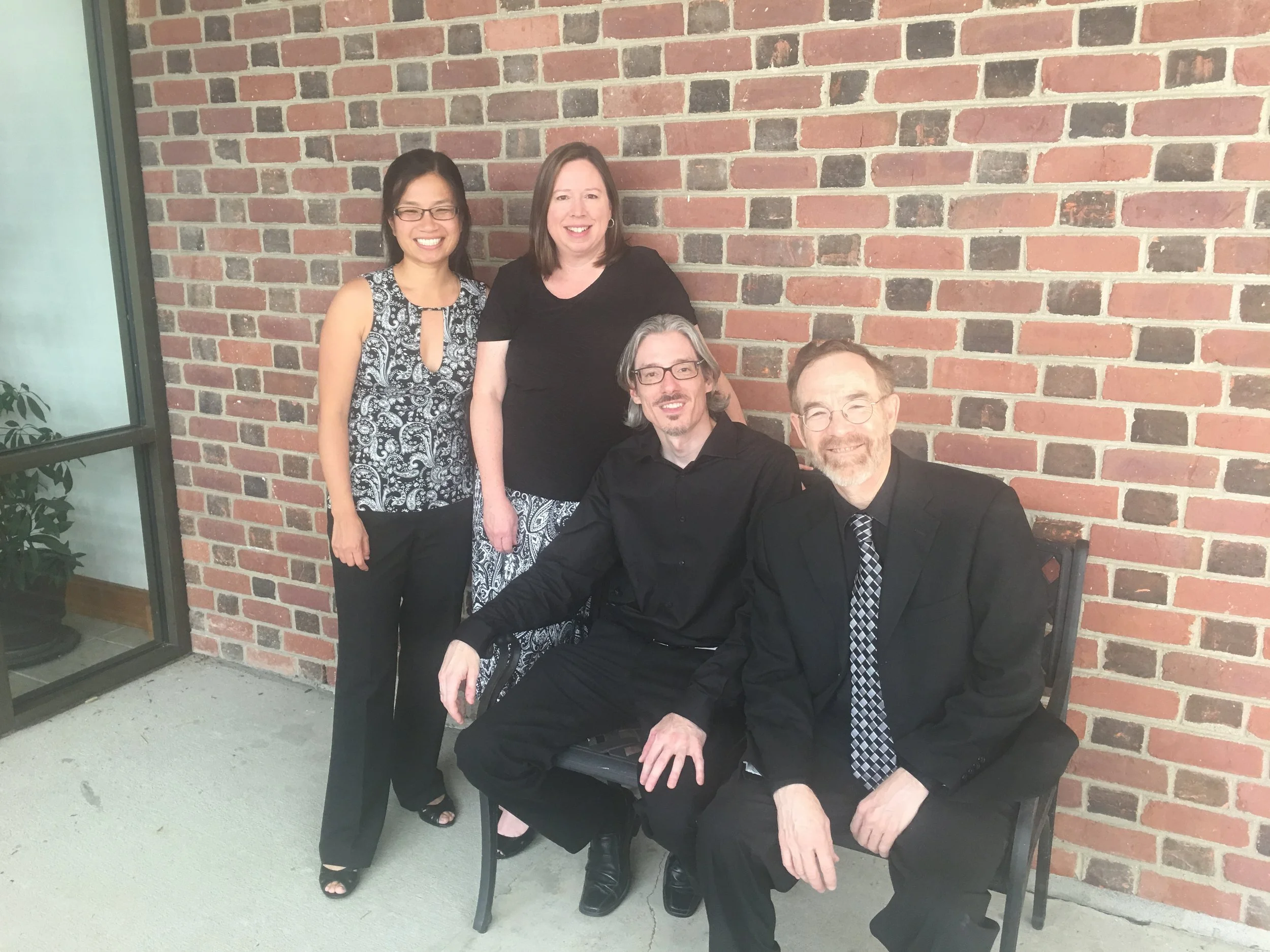 Richmond Chamber Players. Group of four adults, two women and two men, smiling and sitting on a bench outside against a brick wall.