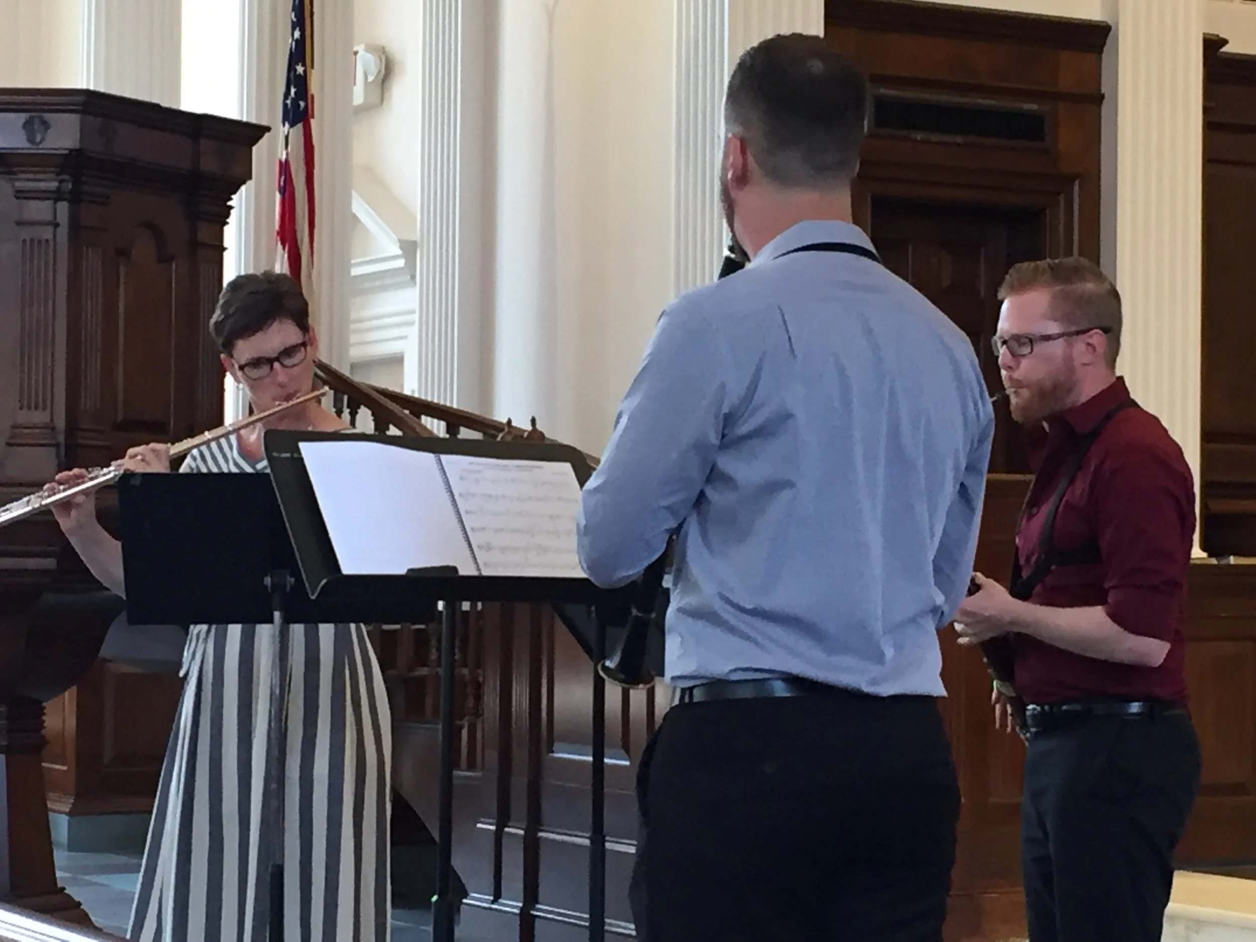 Richmond Chamber Players. Three musicians practicing with music stands in a room with wooden paneling and an American flag in the background.