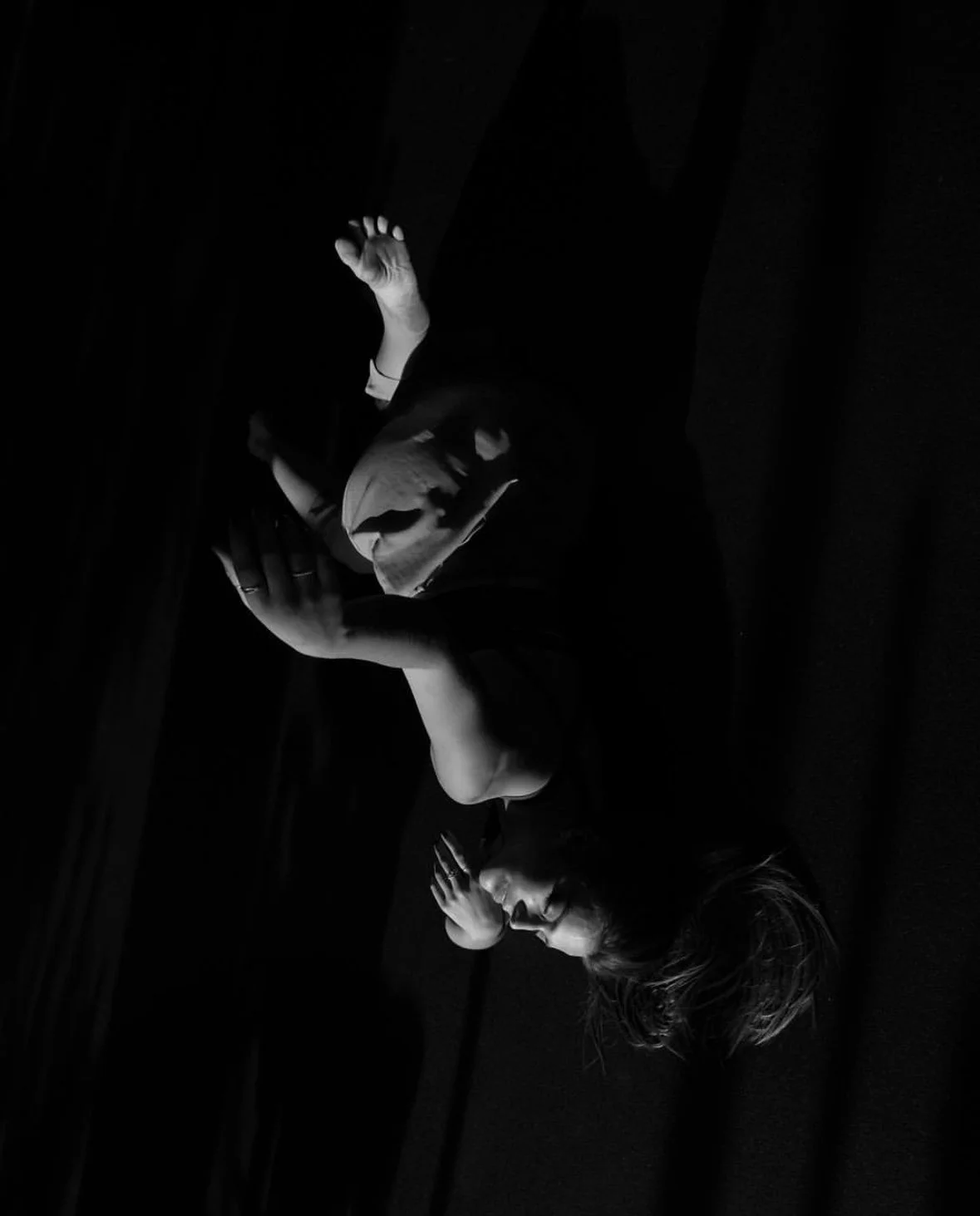 A black and white photo of a woman lying on a surface with her eyes closed, holding a mask to her face with one hand and resting her other hand on her chest.