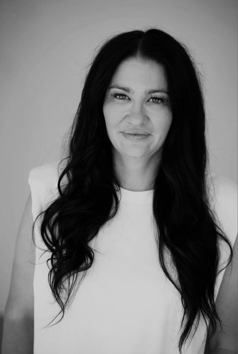 Black and white portrait of a woman with long, wavy dark hair, wearing a sleeveless top, looking directly at the camera with a slight smile.