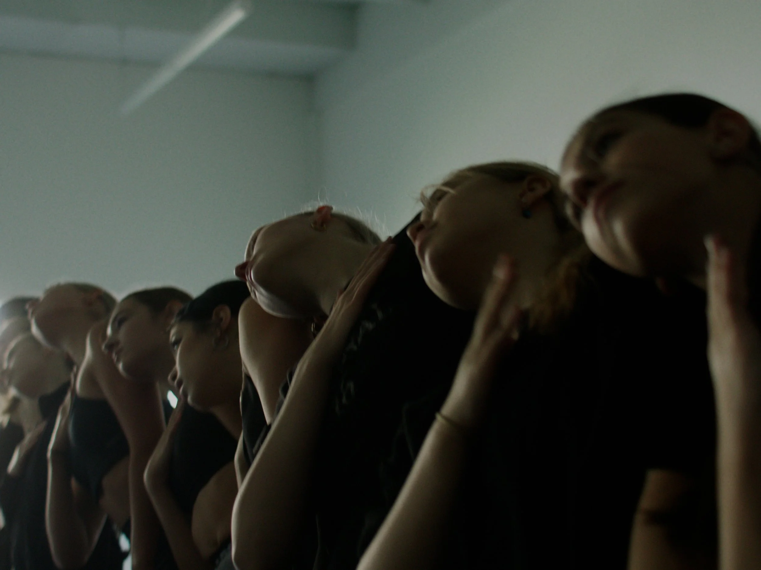 A line of women with attention to their profiles, some touching their necks or faces, indoor setting with plain white wall.