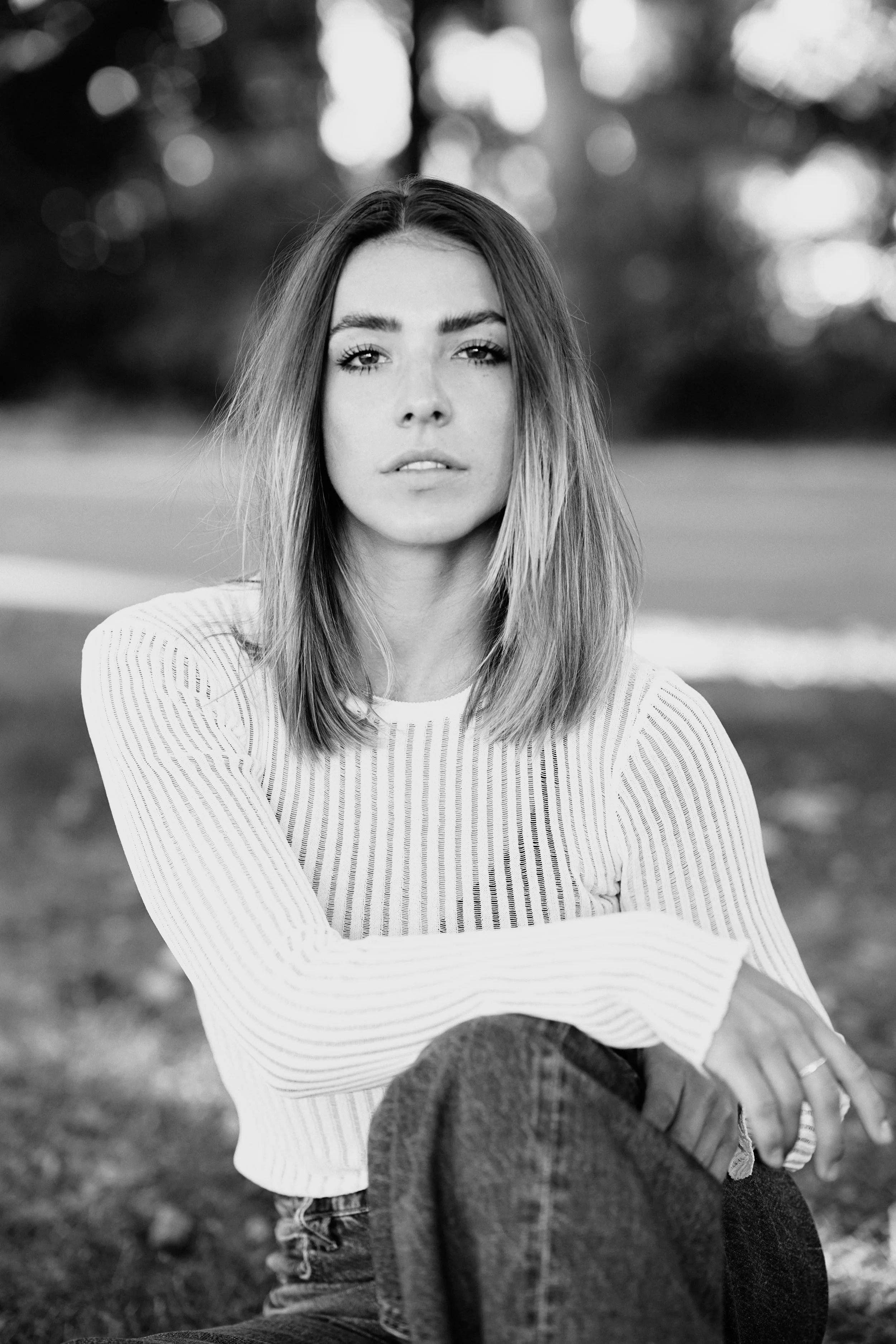 Black and white portrait of a young woman with shoulder-length hair, sitting outdoors on grass, wearing a light striped long-sleeve shirt and jeans, with trees and blurred background behind her.