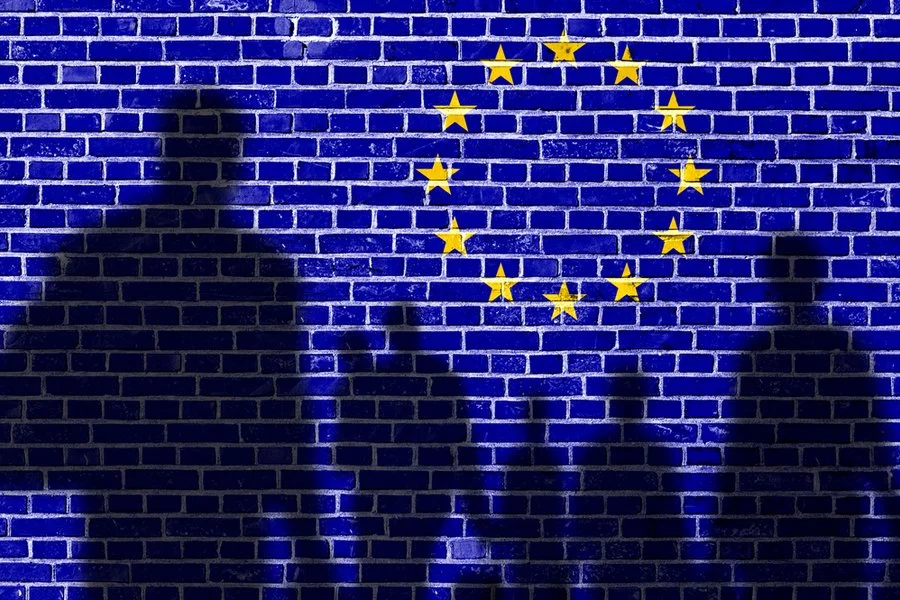 Ombre di due persone e di un bambino proiettate contro un muro di mattoni blu con bandiera dell'Unione Europea dipinta sopra.