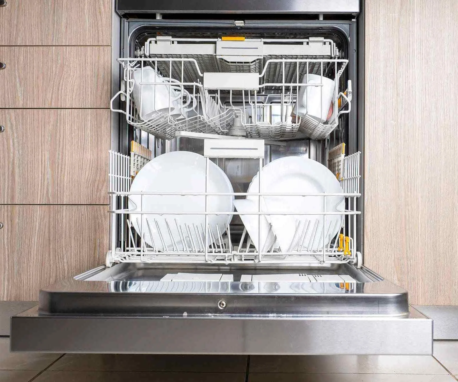 Open built-in dishwasher filled with clean white dishes, plates, bowls, and utensils in a kitchen with wooden cabinets.