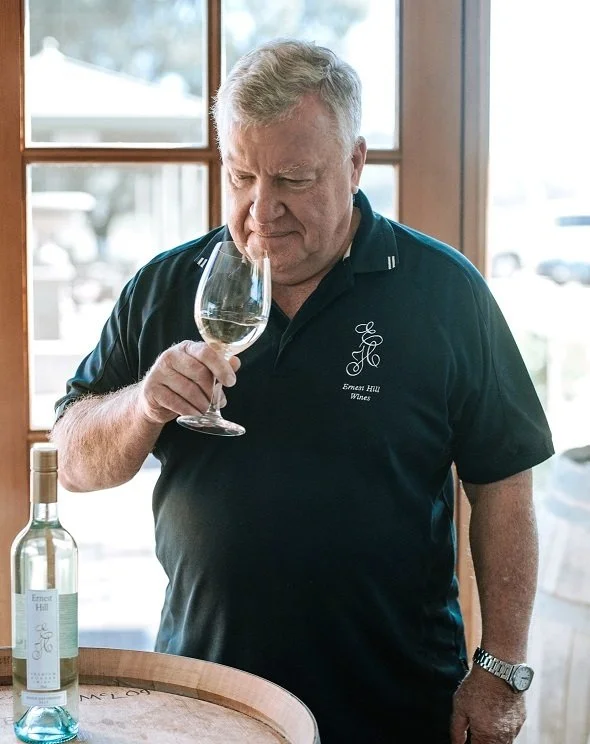 Ross tasting a glass of white wine inside a winery, with a bottle of wine on a barrel nearby.