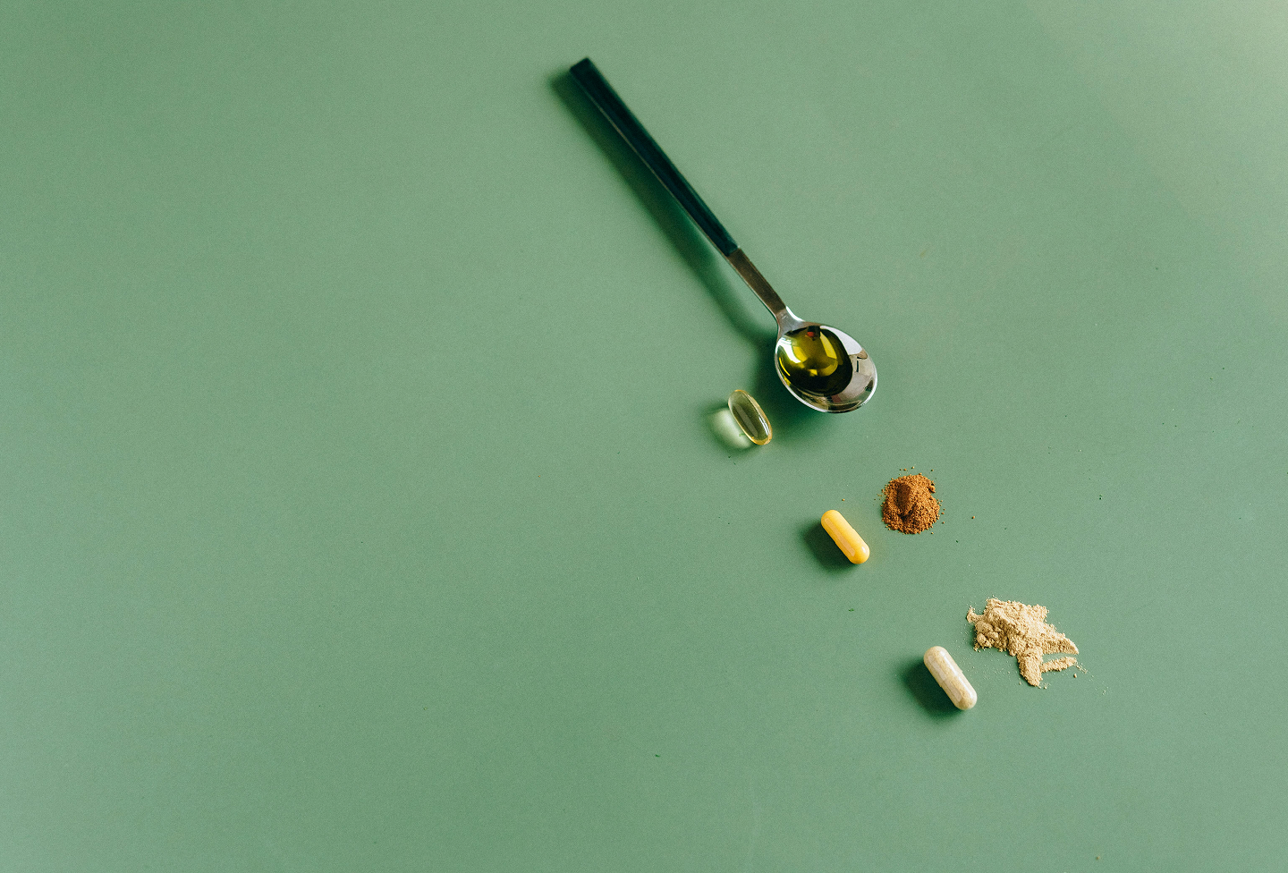Assorted pills, powder, and capsules spread out on a green surface with a spoon containing liquid.