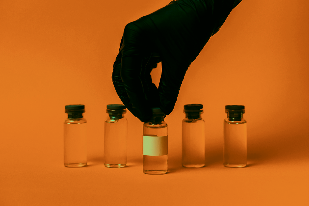 A gloved hand picking up a small glass vial among five similar vials on an orange background.