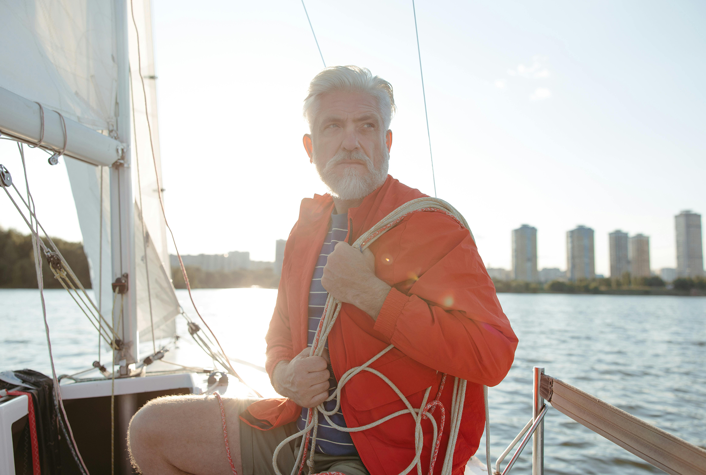 An older man with a gray beard on a sailing boat in a river, carrying ropes, wearing a red jacket and striped shirt, with city buildings in the background, during sunset.