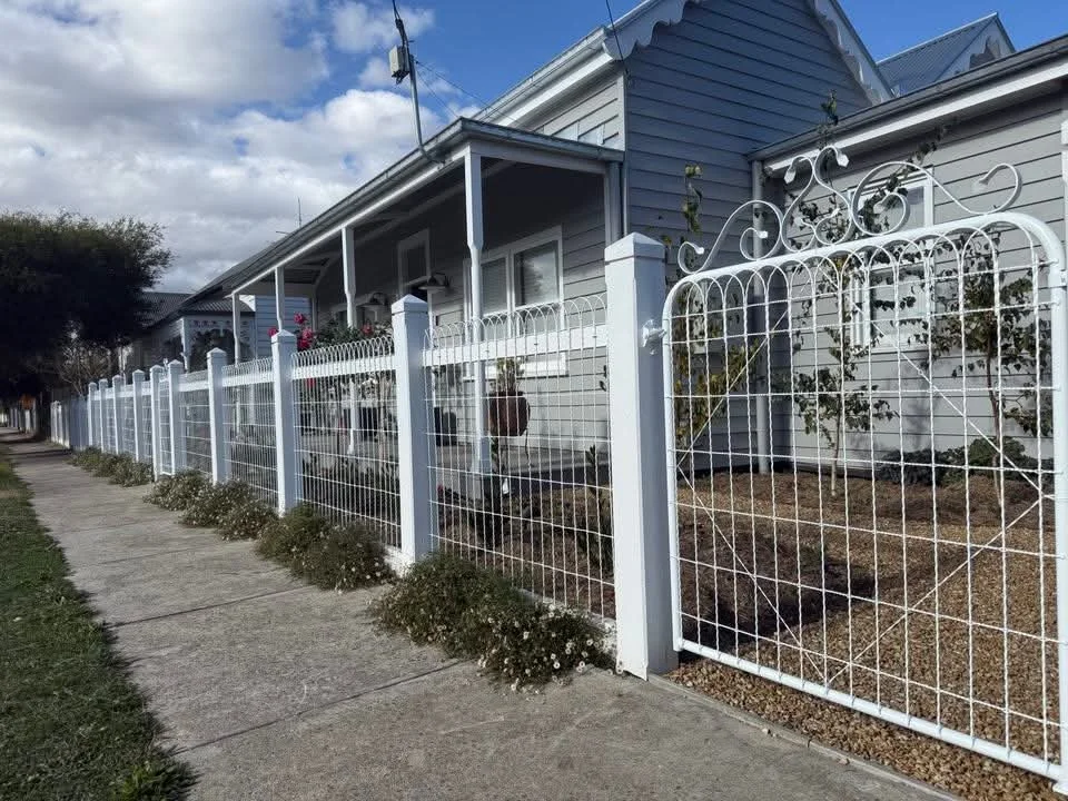 White emu wire fence surrounding a grey house with a front porch, sidewalk in front, and some small bushes along the fence.