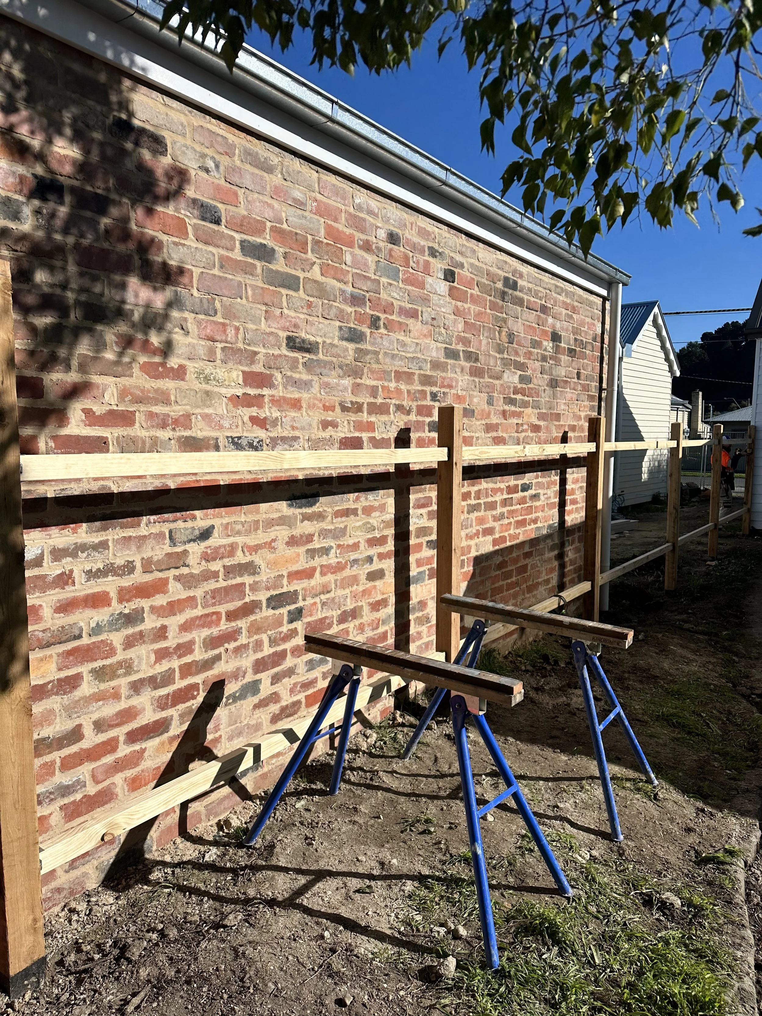 Construction site with a brick wall, wooden fence rails, and sawhorses on the ground.