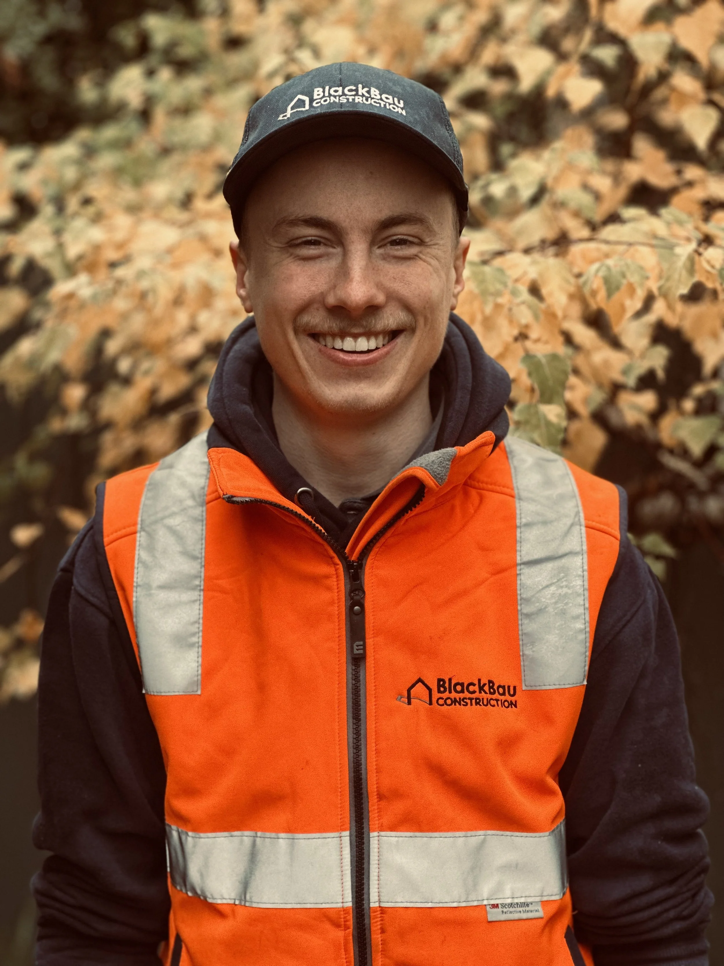 Lucas Black wearing a cap and bright orange safety vest with the BlackBau Construction logo, standing outdoors with autumn leaves in the background.