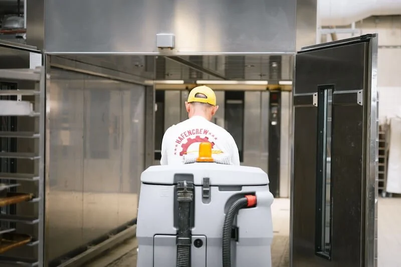 Ein Mann mit gelbem Cap und weißem Shirt in einer industriellen Küche, der einen Reinigungskonvoi benutzt, um den Boden zu reinigen.