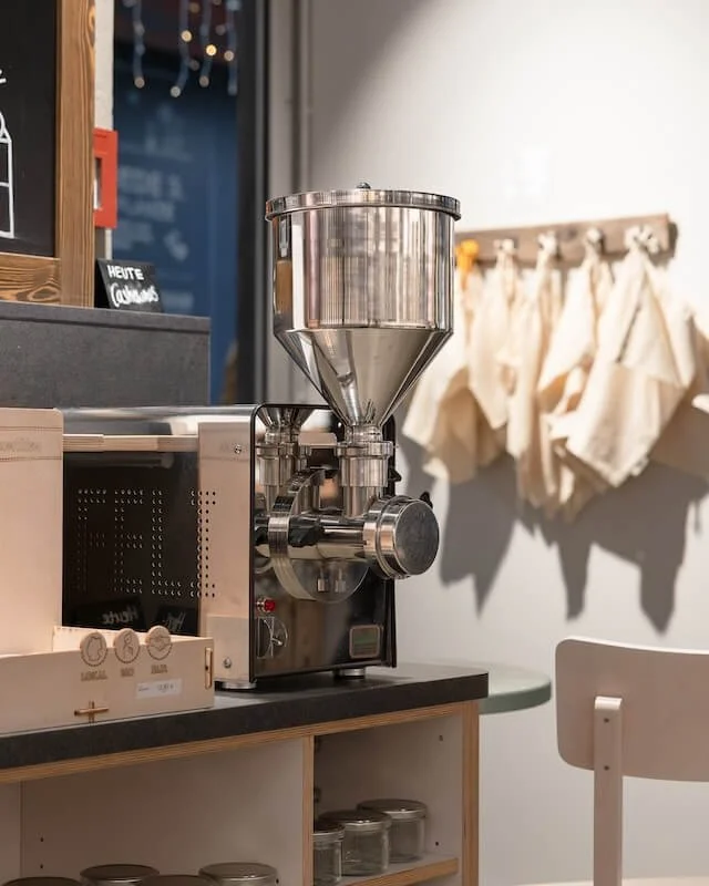 Großes, silbernes Mühlenmaschinen auf einer Laden- oder Café-Theke, im Hintergrund weiße Stofftaschen an der Wand.