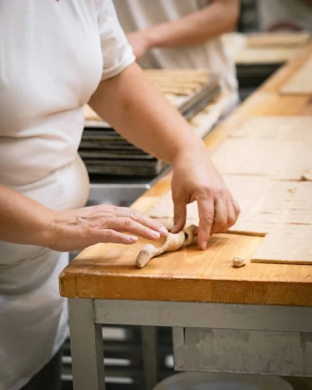 Eine Person, die Teig zu einer langen Wurst formt, auf einem Holzbrett in einer Bäckerei. Auf dem Tisch liegen im Hintergrund mehrere Backbleche mit Teigplatten.