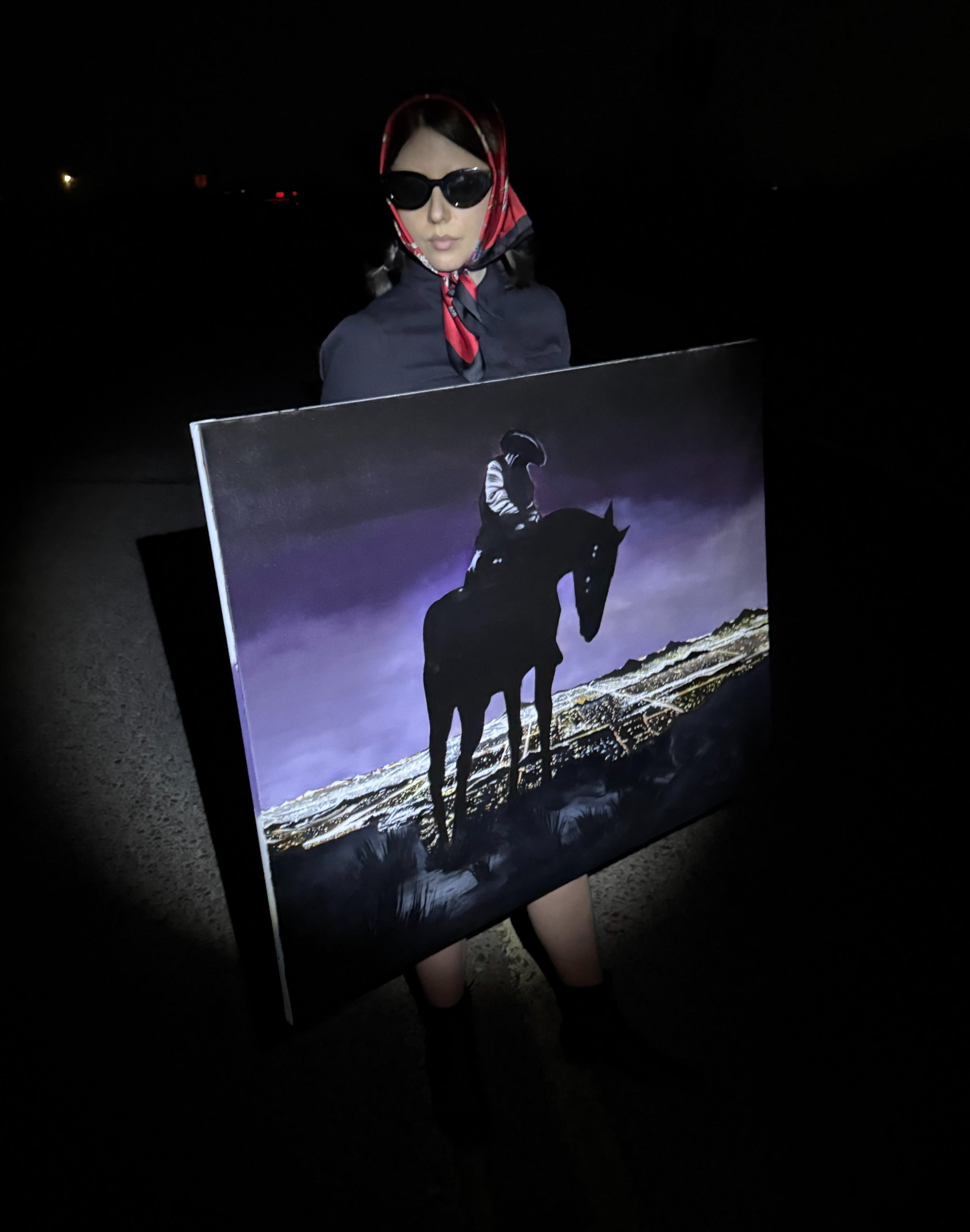 Person wearing sunglasses, a red scarf, a dark jacket, holding a painting of a cowboy on a horse against a landscape at night.